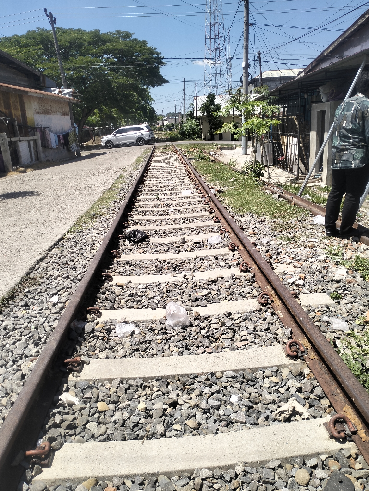 The image depicts a railway track running through a semi-urban area in Lhokseumawe, Indonesia. The tracks are made of rusted metal rails and are laid on concrete sleepers, surrounded by gravel. There is some litter, including plastic bags, scattered around the tracks.
To the left of the tracks, a paved road is visible, with a white SUV parked on it and laundry hanging on a clothesline outside a modest building. Further down the road, a large, leafy tree stands prominently.
To the right of the tracks, there are several buildings, some appearing to be residential with corrugated metal roofs and barred windows. A person wearing a camouflage-patterned shirt and dark pants is standing on the right side of the image, facing away from the camera and towards the railway line. In the background, a tall communication tower is visible against a clear blue sky. The overall impression is one of a functional but somewhat neglected area with a mix of residential and infrastructure elements.