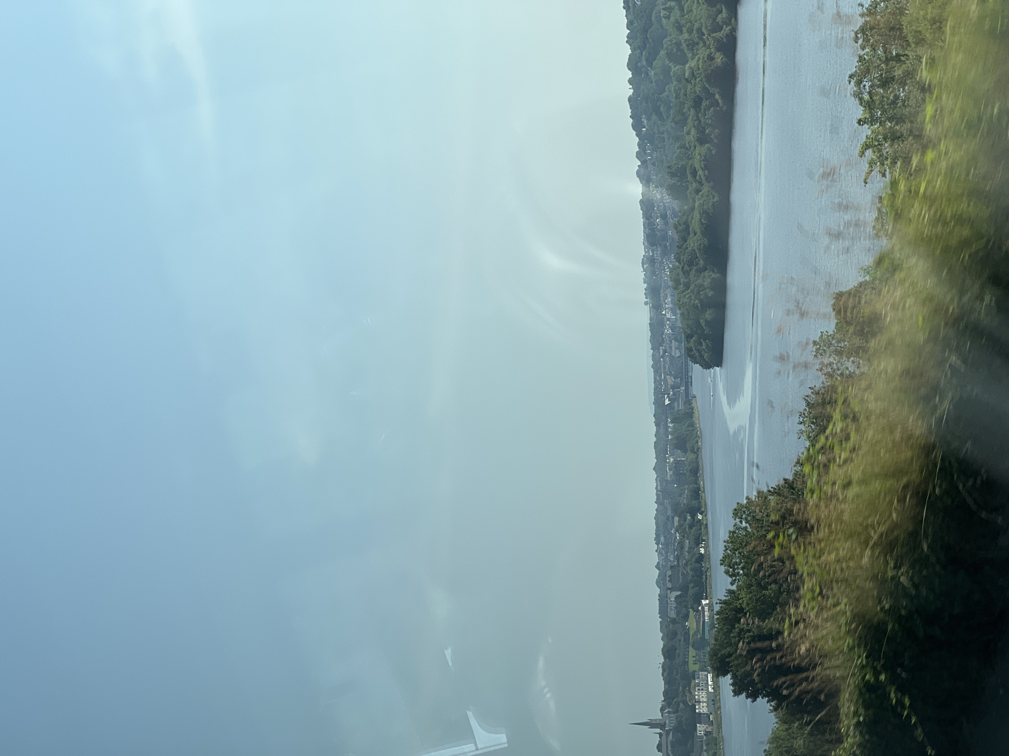 The image shows a wide river, likely the River Foyle, in the city of Derry/Londonderry, United Kingdom. On the left bank of the river, there is a view of a cityscape with several buildings, including a prominent church spire. The cityscape is nestled amongst lush green trees. The river itself is calm and reflects the light blue sky. On the right bank of the river, there is a dense area of trees. The foreground of the image is blurred, suggesting it was taken from a moving vehicle, possibly a train or car. The sky is mostly clear with some faint wispy clouds and some reflections on the window. The overall impression is a scenic view of a river passing through a city surrounded by nature.