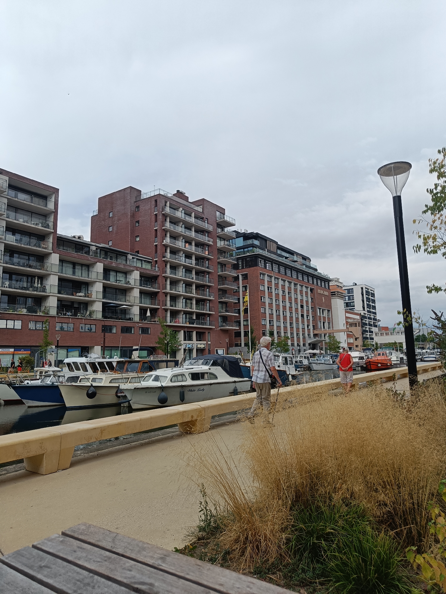 The image depicts a waterfront scene in the city of Leuven, Belgium, likely along the Vaart, given the presence of docked boats and modern brick buildings. In the foreground, a paved walkway runs alongside a canal where several boats are moored. A light-colored, modern bench is visible to the left. Two individuals are walking on the walkway: an older man with gray hair, wearing a checkered shirt and light-colored trousers, carrying a walking stick, and a woman with red hair wearing a red top and white shorts. Beyond the walkway, a line of boats, including a white one labeled "White Lady" and another named "CASTOR," are moored. Behind the boats, a series of multi-story brick buildings with numerous balconies and large windows dominate the background. A prominent lamppost stands on the right side of the walkway. The sky is overcast and gray. The overall environment suggests a commercial or residential development along a waterway.