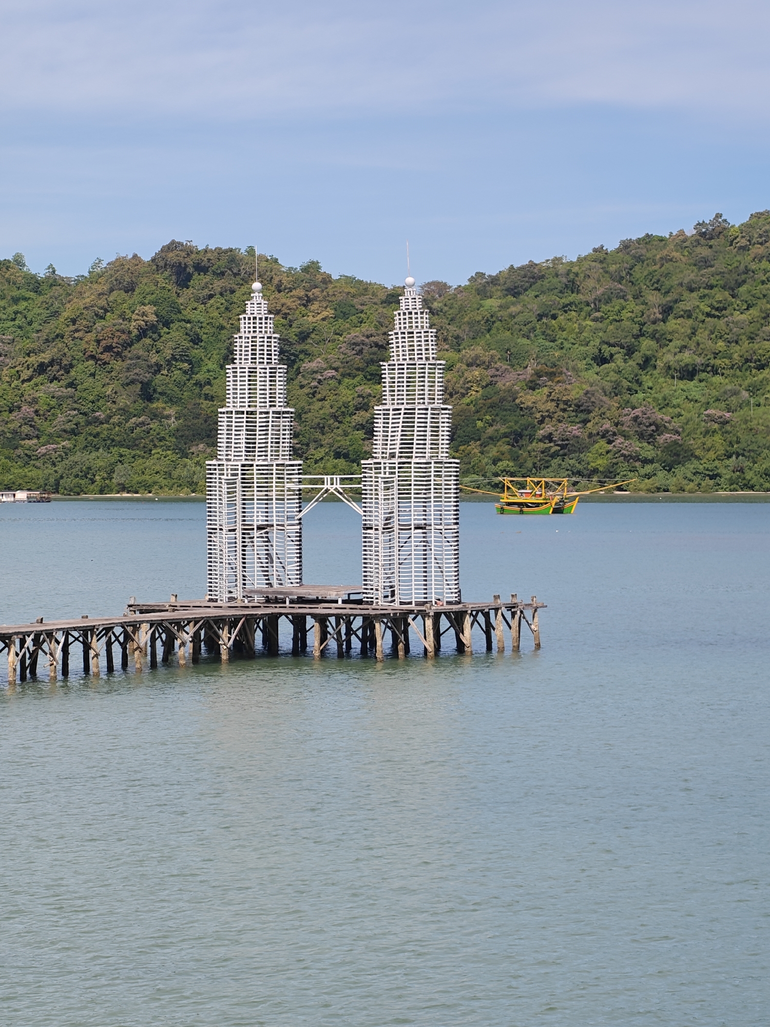 The image shows a replica of the Petronas Twin Towers made of white wooden planks, standing on a wooden pier in the water. The replica is located in Layeun, Indonesia. A boat with a yellow and green body is visible on the water behind the replica. A wooded shoreline is in the background. The sky is blue.