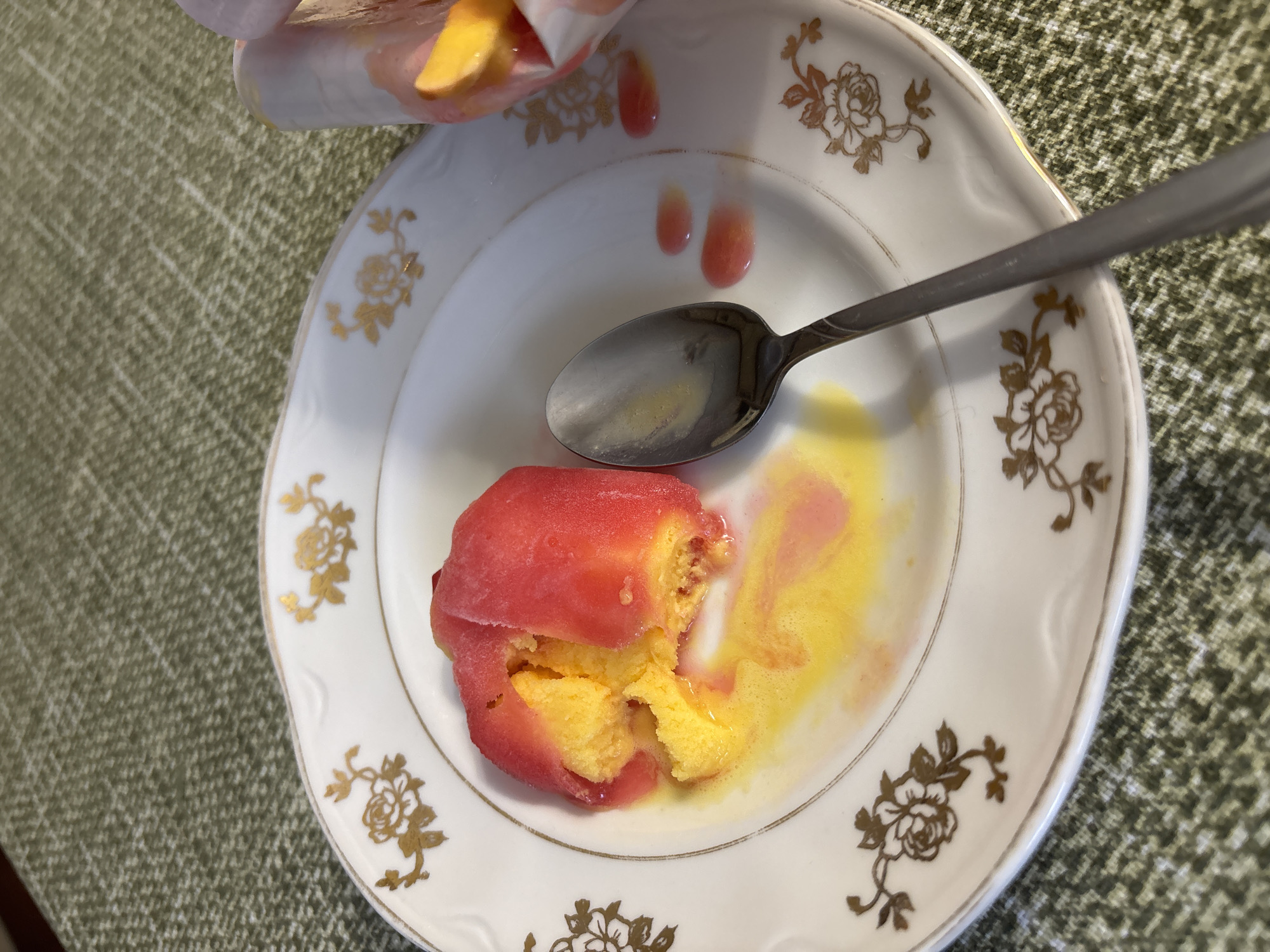 An overhead shot on a textured green tablecloth shows a white plate with a golden floral pattern. A melting ice cream confection, with a red outer shell and a yellow interior, sits on the plate. A silver spoon rests on the plate next to the ice cream, and there are drips of red and yellow liquid on the plate. In the upper right corner, a partially visible pink wrapper holds slices of what appears to be fruit, possibly peaches or apricots. The image is taken in Sinyavino, Russia.