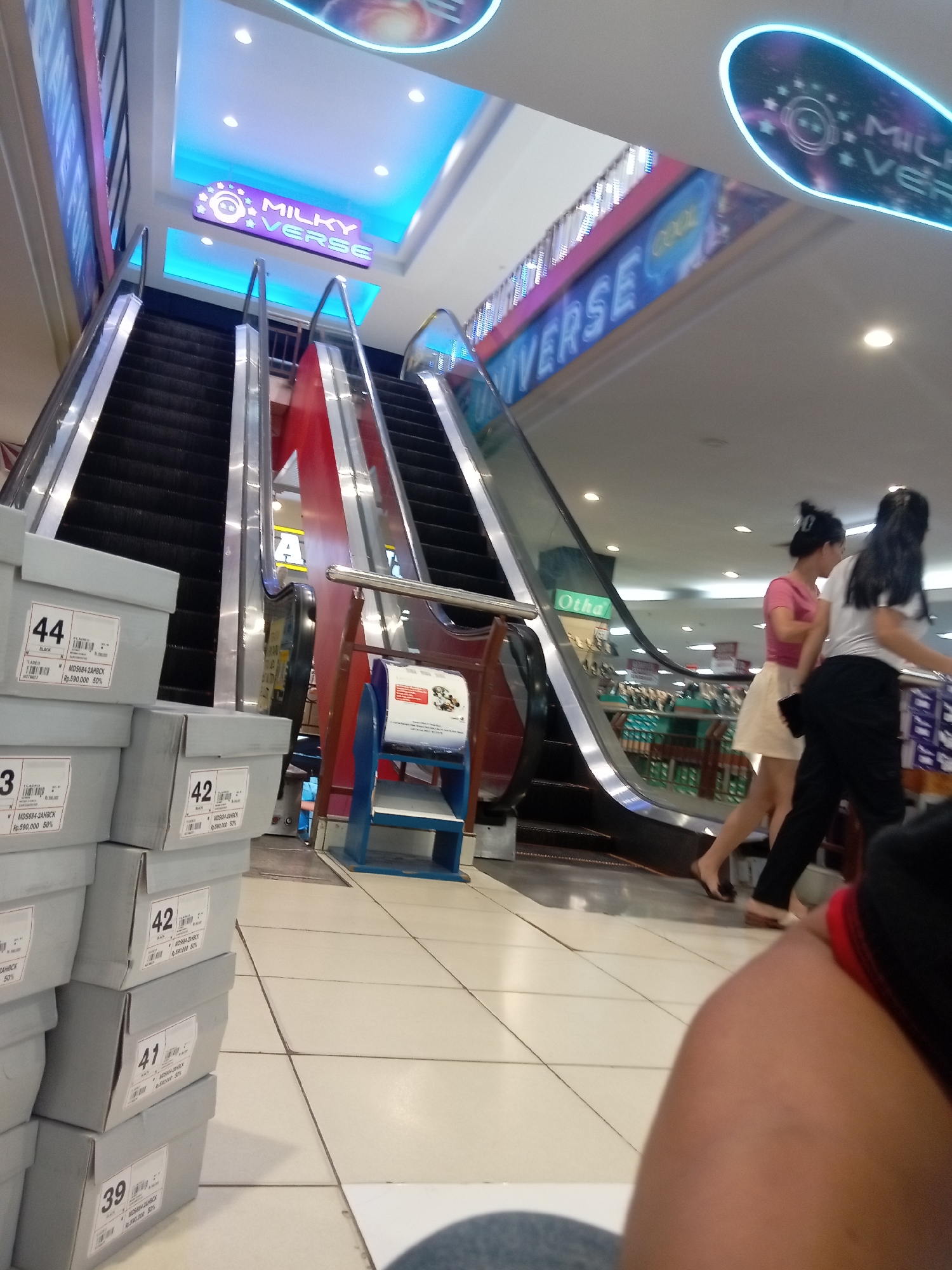 This is a street-level, eye-level medium shot taken in a shopping mall in Batam, Indonesia, depicting an escalator area. The image shows two escalators, one ascending and one descending, with metallic handrails and black steps. Above the escalators, a blue illuminated ceiling is visible, featuring a purple sign with the text "MILKY VERSE" in white lettering. To the right of the escalators, two women are walking away from the camera. The woman on the left wears a pink top and a light-colored skirt, while the woman on the right wears a white t-shirt and black pants. In the foreground, stacks of shoe boxes are piled up. Some boxes have labels with numbers such as "44", "43", "42", "41", and "39". The floor is tiled in a beige color. The overall environment suggests a commercial retail space within a shopping center.
