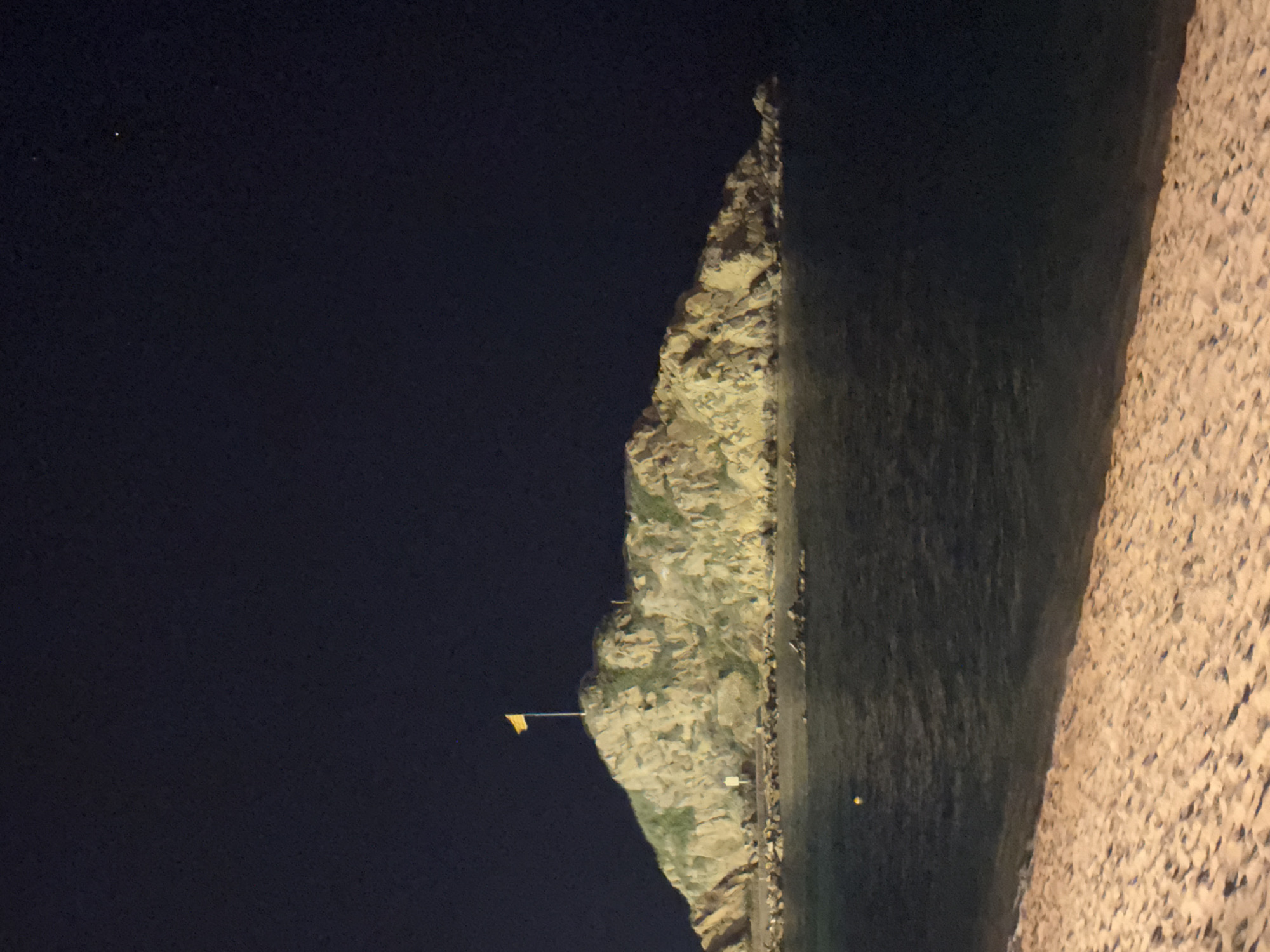 The image depicts a nighttime view of Sa Palomera, a rocky promontory located in Blanes, Spain. The promontory is topped with vegetation and a yellow flag flying from a flagpole. The sea is calm and dark, with subtle ripples reflecting the ambient light. In the foreground, a sandy beach stretches out, illuminated by artificial lights, showing the texture of the sand and the gentle slope towards the water. The sky is dark and clear, with a few distant stars visible. There are no people clearly discernible in the image, though faint human figures may be present near the base of the promontory. The scene is a serene and natural landscape illuminated by artificial light sources, highlighting the contours of the land and the stillness of the night.