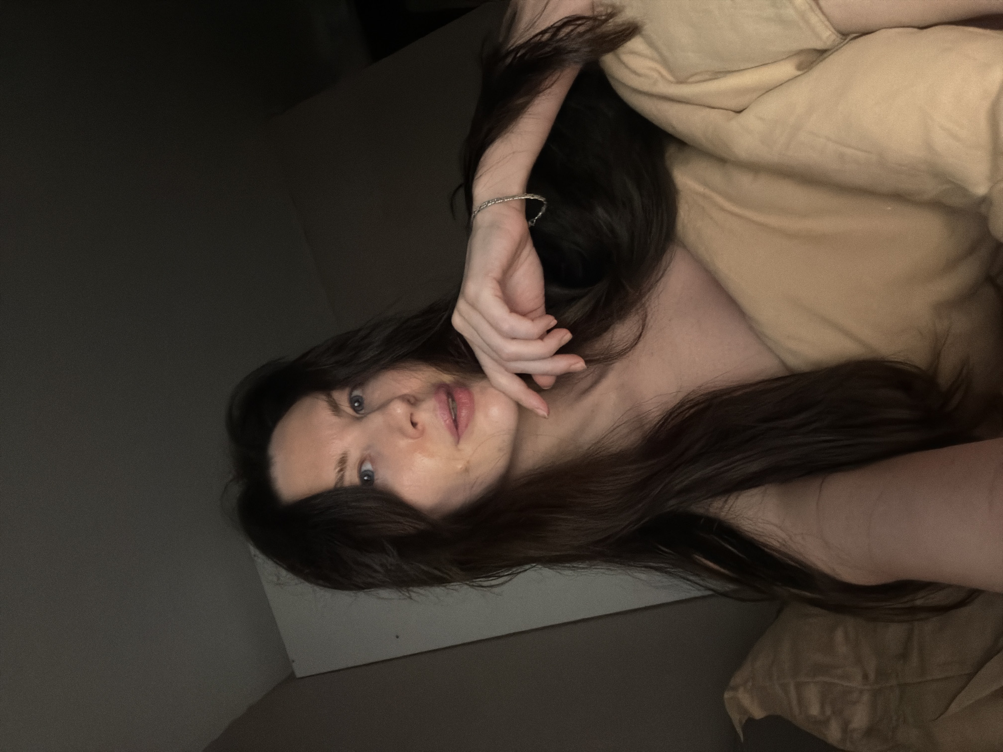 A pale-skinned woman with long, dark hair lies in bed under a beige blanket. Her face is visible, with blue eyes and her hand resting near her face. She is wearing a silver bracelet on her wrist. The background is a dimly lit room with a gray wall. The location is unknown and it is not possible to determine the city of Perm, Russia.