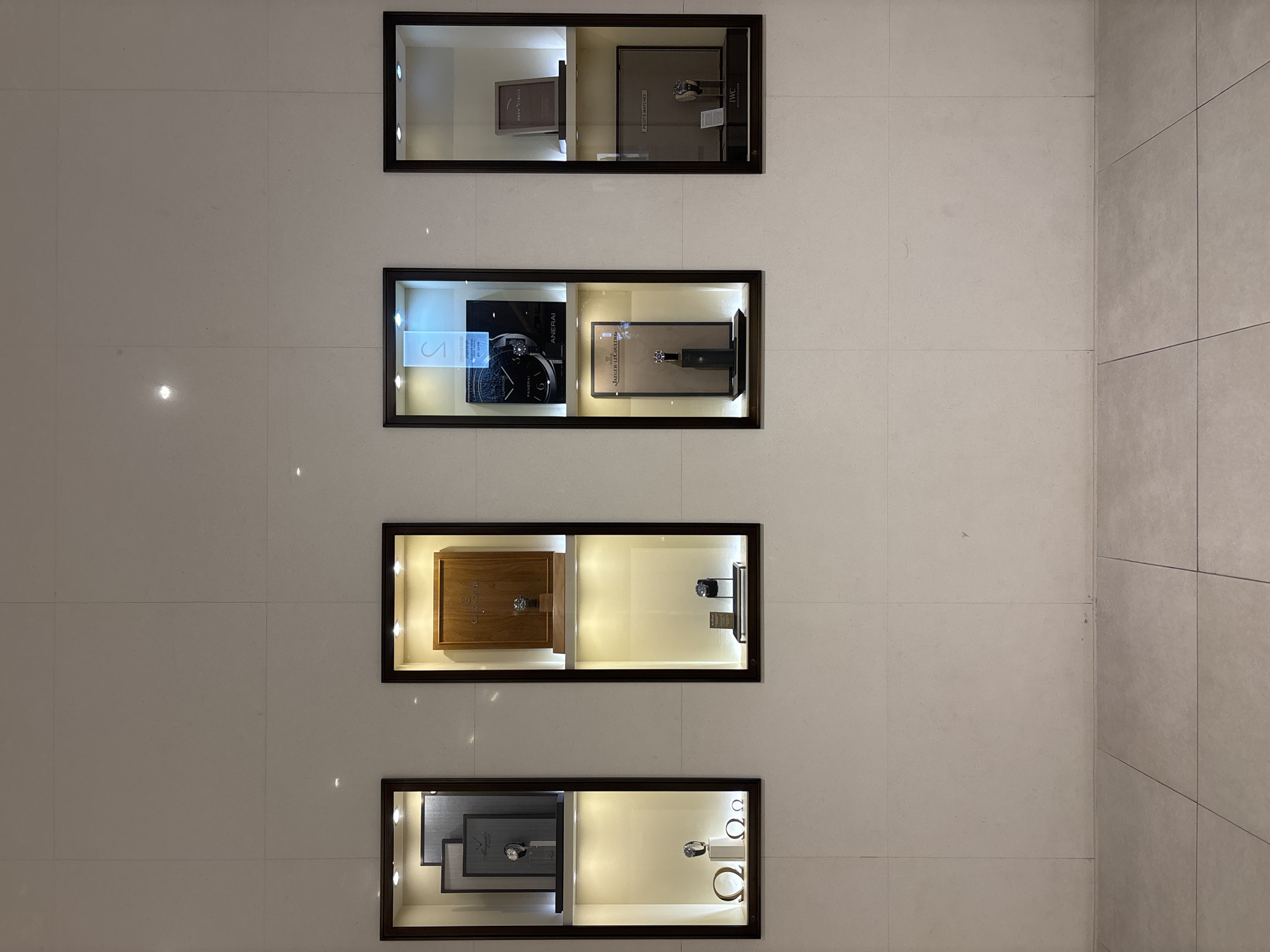 The image shows a retail display wall with four illuminated glass-fronted cabinets, each displaying luxury watches and related accessories. The cabinets are arranged horizontally on a white tiled wall. From left to right, the first cabinet contains several framed items and a watch on a stand, as well as three omega symbols. The second cabinet features a wooden box with a watch inside, and below it, another watch on a stand. The third cabinet displays a boxed watch and a watch on a stand from Jaeger-LeCoultre. The fourth cabinet showcases a framed picture, a boxed set of "PILOT'S WATCHES," and an IWC Schaffhausen watch. The floor is tiled with a light grey material. The scene is likely within a watch boutique or a luxury department store. This specific display is located in the luxury watch section of the Shinsegae Centum City department store in Busan, South Korea.