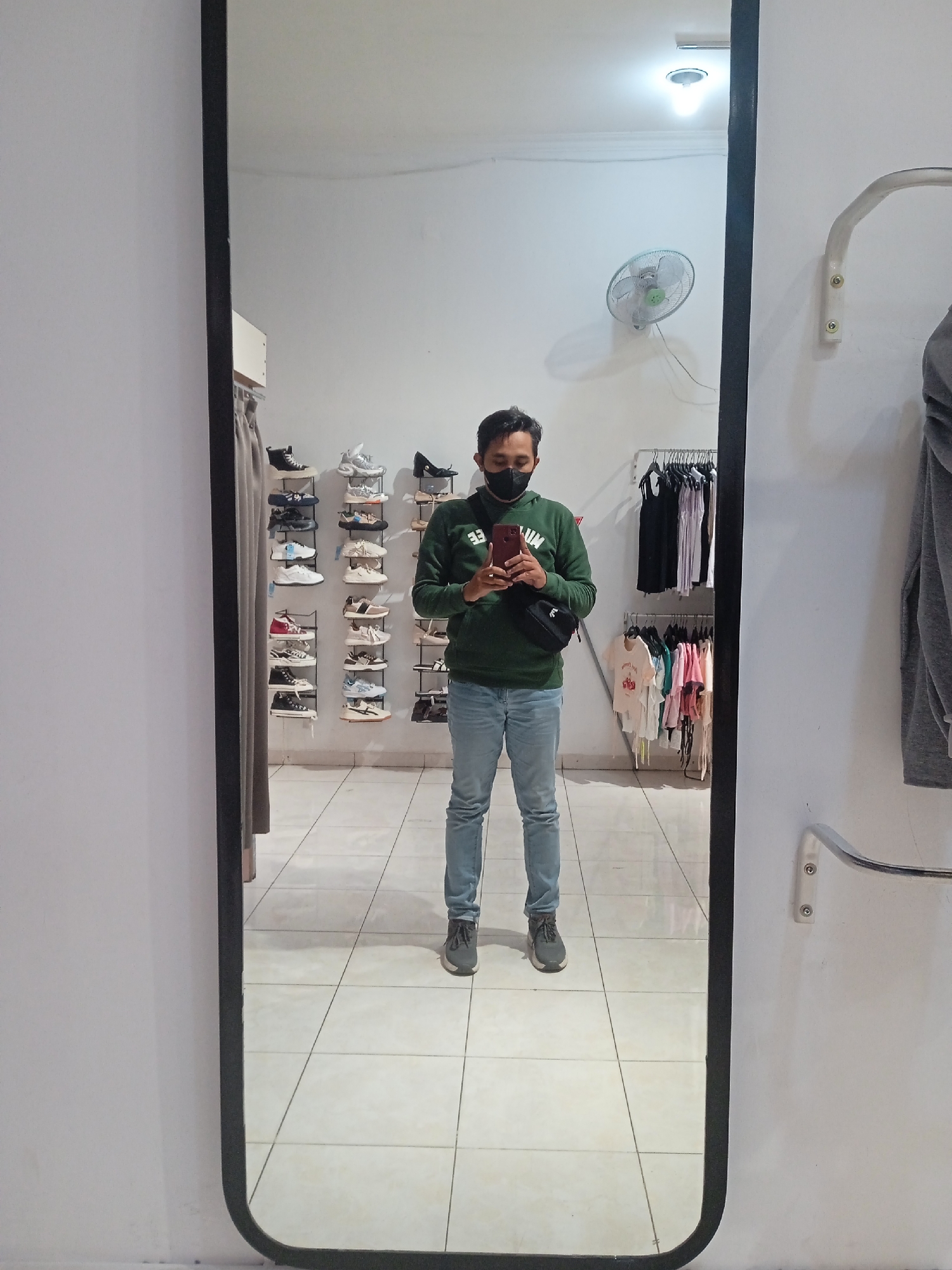 A full-body mirror reflects a young man standing in what appears to be a retail store or a dressing room in Semarang, Indonesia. He is wearing a dark green hooded sweatshirt, blue jeans, a black face mask, and dark sneakers. A black crossbody bag is slung across his chest. He is holding a red smartphone in his right hand, seemingly taking the selfie.
The background of the reflection shows shelves filled with various shoes, ranging from sneakers to boots, arranged neatly on white walls. Further back, clothing racks with a variety of apparel are visible, including t-shirts and dresses. A white oscillating fan is mounted on the wall above the shelves of shoes. The floor is tiled with a pattern of light-colored squares. The lighting in the room is bright, with an overhead fluorescent light visible on the ceiling. The overall impression is of a small retail space, possibly a shoe or clothing boutique.