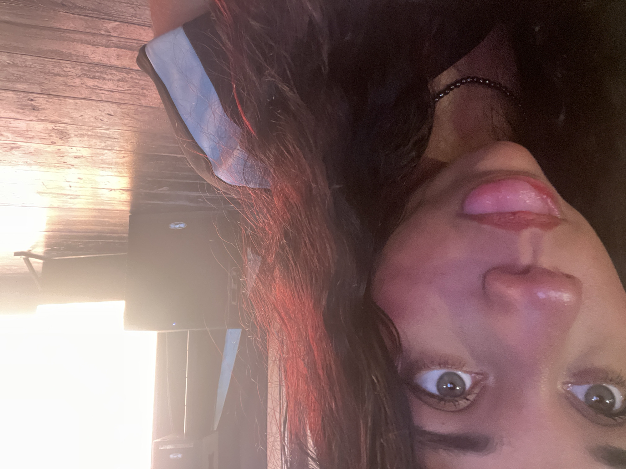 The image is a close-up, selfie-style photograph of a young woman's face and upper body, taken indoors. The woman has dark hair with reddish highlights, green eyes, and her tongue is extended. She is wearing a dark top with blue and white stripes on the sleeve. The background appears to be a stage or performance area, with large speakers and a bright, illuminated screen or light source. The floor is made of wooden planks. The lighting is warm and slightly dramatic, casting some reddish tones on the woman's hair and skin. The overall impression is of a casual, playful moment captured during an event. There is no information in the image to indicate a specific location in Porlamar, Venezuela.