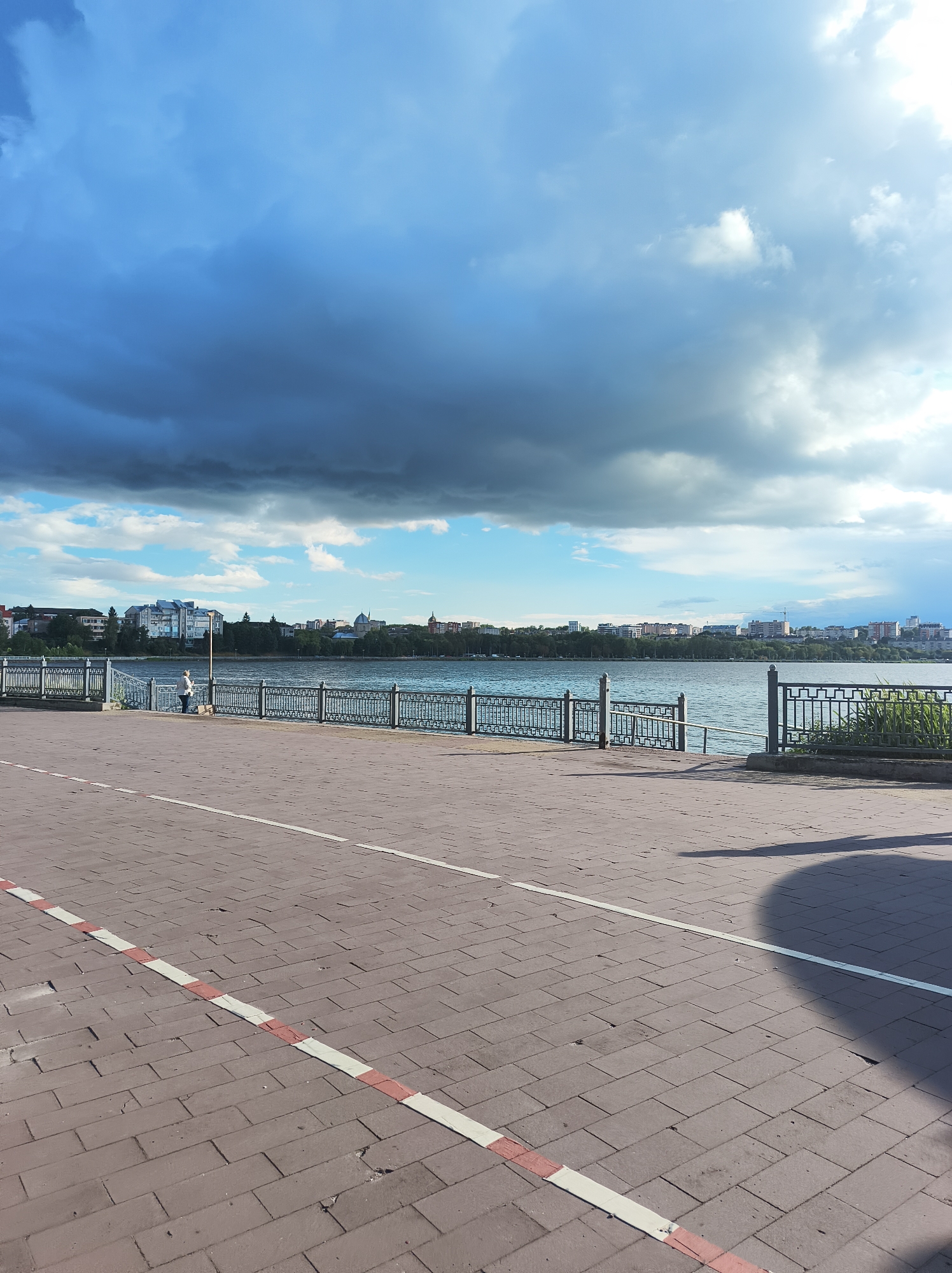 The image captures a waterfront promenade in Ternopil, Ukraine, with a focus on the Ternopil Lake. In the foreground, a paved walkway made of reddish-brown bricks is visible, marked by a white line and a red and white striped line, indicating a pedestrian path or bike lane. A dark shadow, likely cast by a person or object, falls across the walkway from the right.
In the mid-ground, a black metal fence with an ornate design separates the promenade from the Ternopil Lake. A single individual, appearing to be a woman wearing a light-colored top and dark pants, stands by the fence, facing the water. She is accompanied by a small, dark object on the ground beside her, possibly a bag.
The Ternopil Lake stretches across the horizon, its surface rippled by a gentle breeze. On the far bank of the lake, the cityscape of Ternopil unfolds, featuring a mix of residential buildings, some with modern architecture and others with older designs. Trees line the waterfront, and further in the distance, more buildings and structures are visible, including what appears to be a church dome.
The sky above is dramatic, with a significant portion occupied by dark, heavy clouds that suggest an approaching storm or recent rainfall. These dark clouds are contrasted by patches of brighter blue sky and lighter, wispy clouds, creating a dynamic and somewhat moody atmosphere. The overall impression is that of a public space by a large body of water, with elements of urban life and natural weather phenomena.