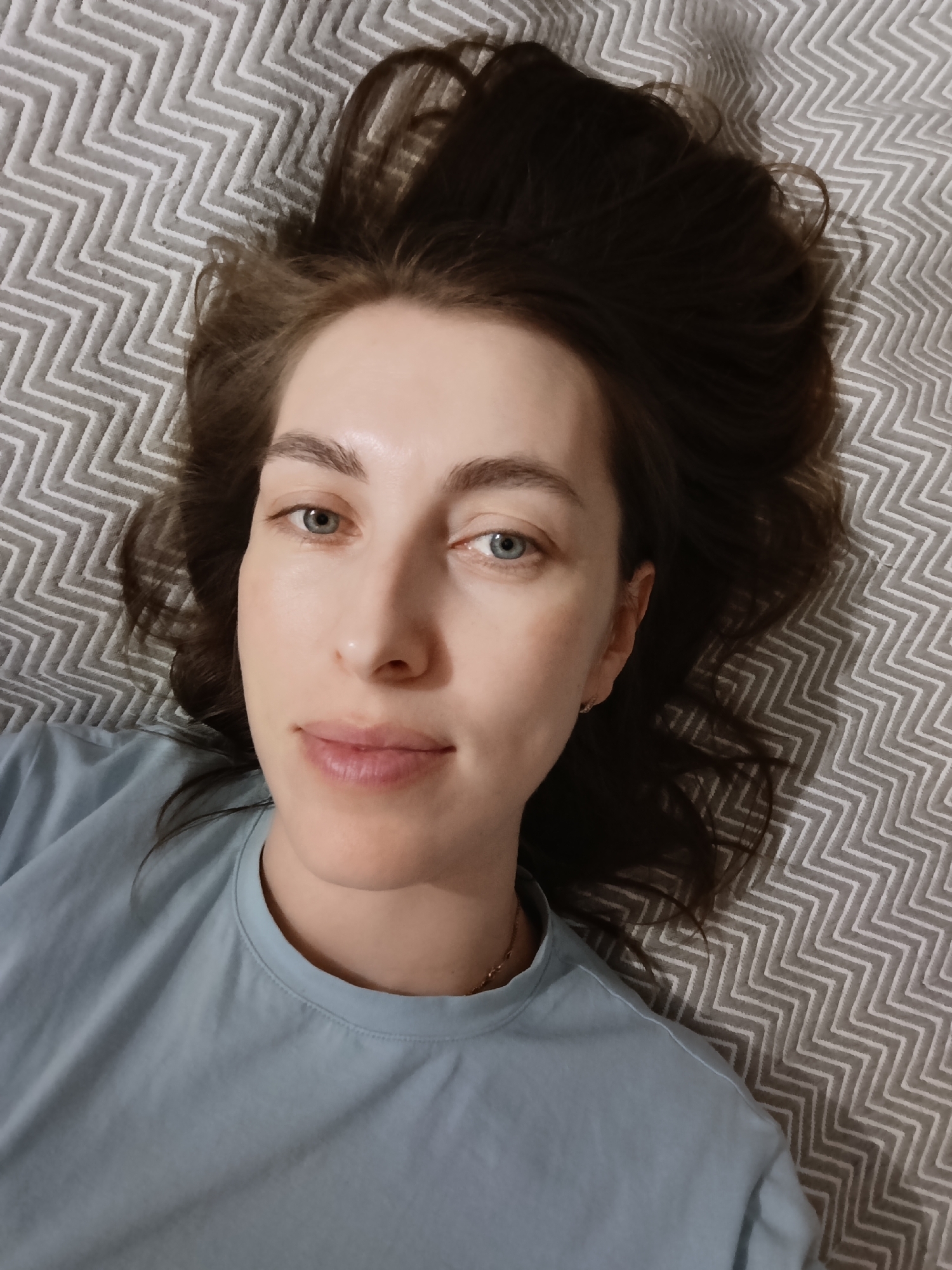 A woman with fair skin, blue eyes, and brown hair is lying on a gray and white chevron-patterned surface, possibly a bed. She is wearing a light blue t-shirt and a thin gold necklace. The image is a close-up shot of her face and upper body. The location is unknown, and there is no indication of it being Chaikovsky, Russia, in the image.