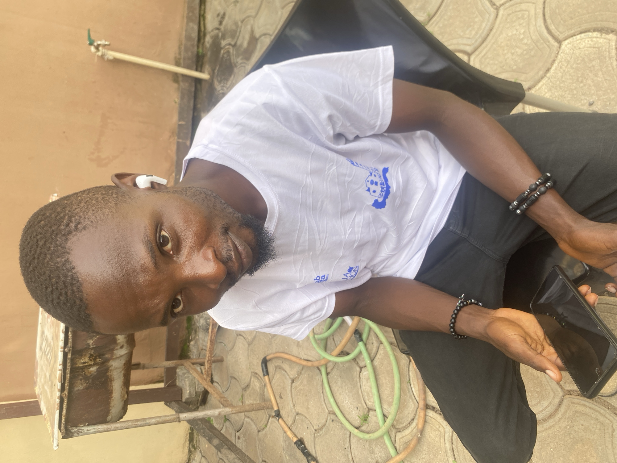 A Black man wearing a white t-shirt and black pants is seated outdoors on a black chair. He has short, dark hair and a goatee. He is wearing white earbuds and two beaded bracelets on his left wrist. The man is holding a black smartphone in his left hand. In the background, there is a rusty metal container on a stand, a green and orange hose, a faucet with a pipe, and a tan wall. The ground is paved with interlocking concrete blocks. The scene appears to be in Barden Yana, Nigeria.
