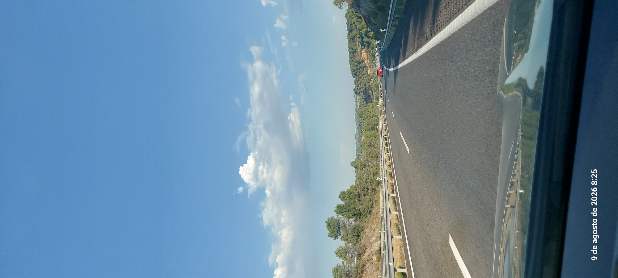 The image depicts a road scene, likely a highway, captured from a vehicle's perspective. The road is asphalt with clear white lane markings. Guardrails line the sides of the road, separating it from the adjacent landscape. The landscape consists of rolling hills covered with dense green trees, possibly pine trees, and rocky terrain on the higher slopes. The sky is a vibrant blue with scattered white clouds. A single red car is visible on the road ahead, driving in the same direction. The bottom of the image shows the hood of the vehicle from which the photo was taken. A timestamp in the lower left corner reads "9 de agosto de 2026 8:25". This suggests the location is Vinaixa, Spain, based on the date and the common style of highway and landscape seen in that region. The overall scene conveys a sense of travel on a clear day.