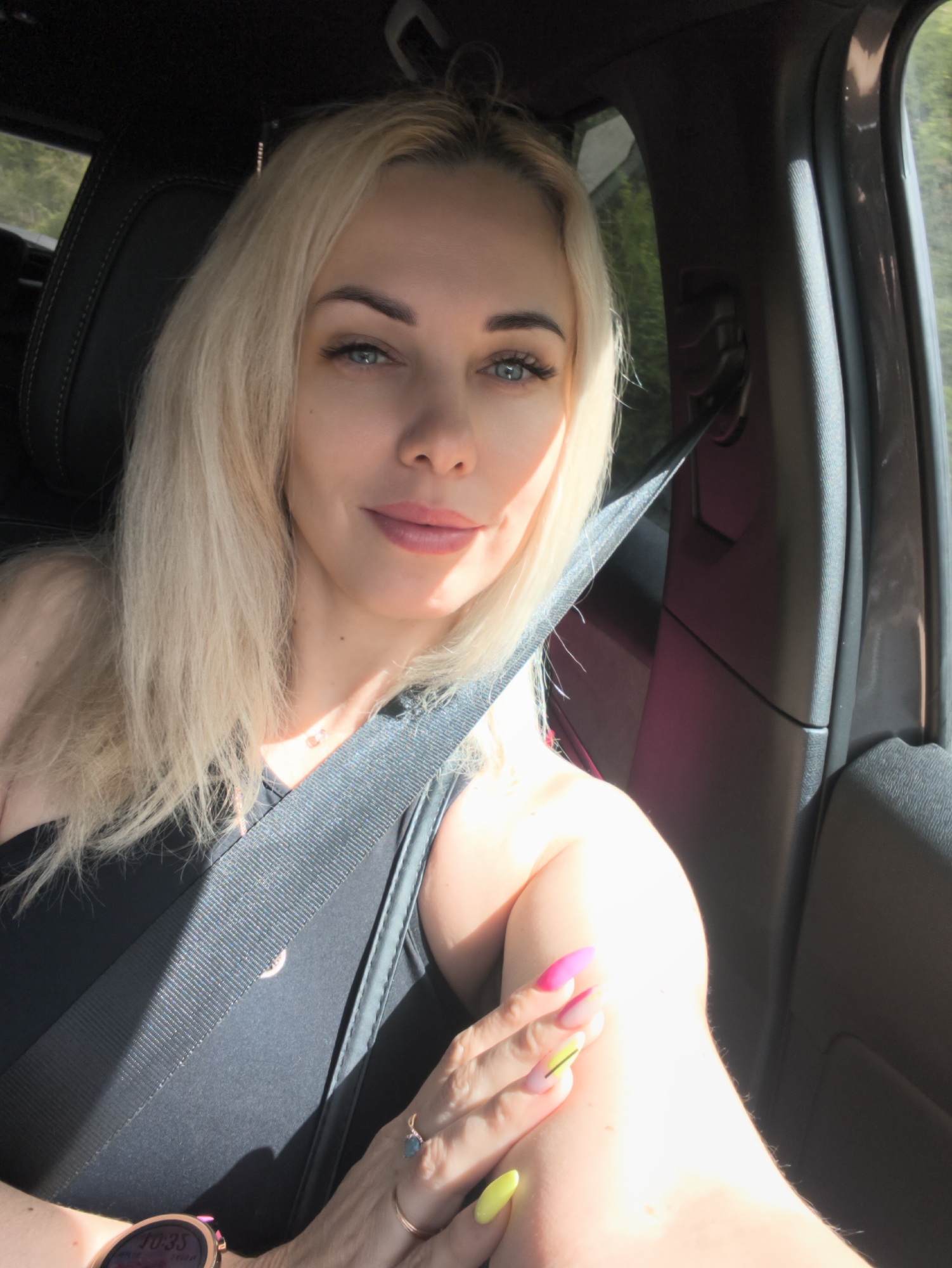 A woman with blonde hair and blue eyes is sitting in the driver's seat of a car, wearing a black tank top and a seatbelt. She has light skin and is wearing a watch on her left wrist and rings on her fingers. Her nails are painted in various bright colors. The interior of the car is visible, including the seat, window, and door. The location cannot be determined as being within Kamchatka, Russia from the image.