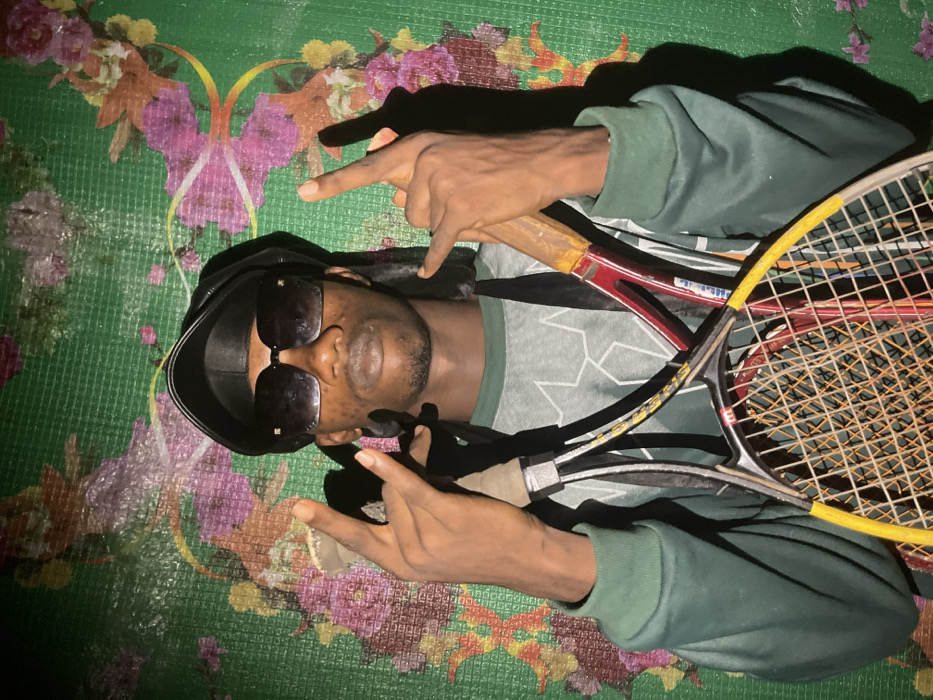 A person lies on a green patterned mat, wearing sunglasses and a head covering. They hold two tennis rackets. The person is dressed in a green and gray patterned top with green sleeves. They are making a hand gesture with both hands, extending their index and middle fingers. The background mat has a floral design with pink and purple elements. The overall lighting of the image is subdued.