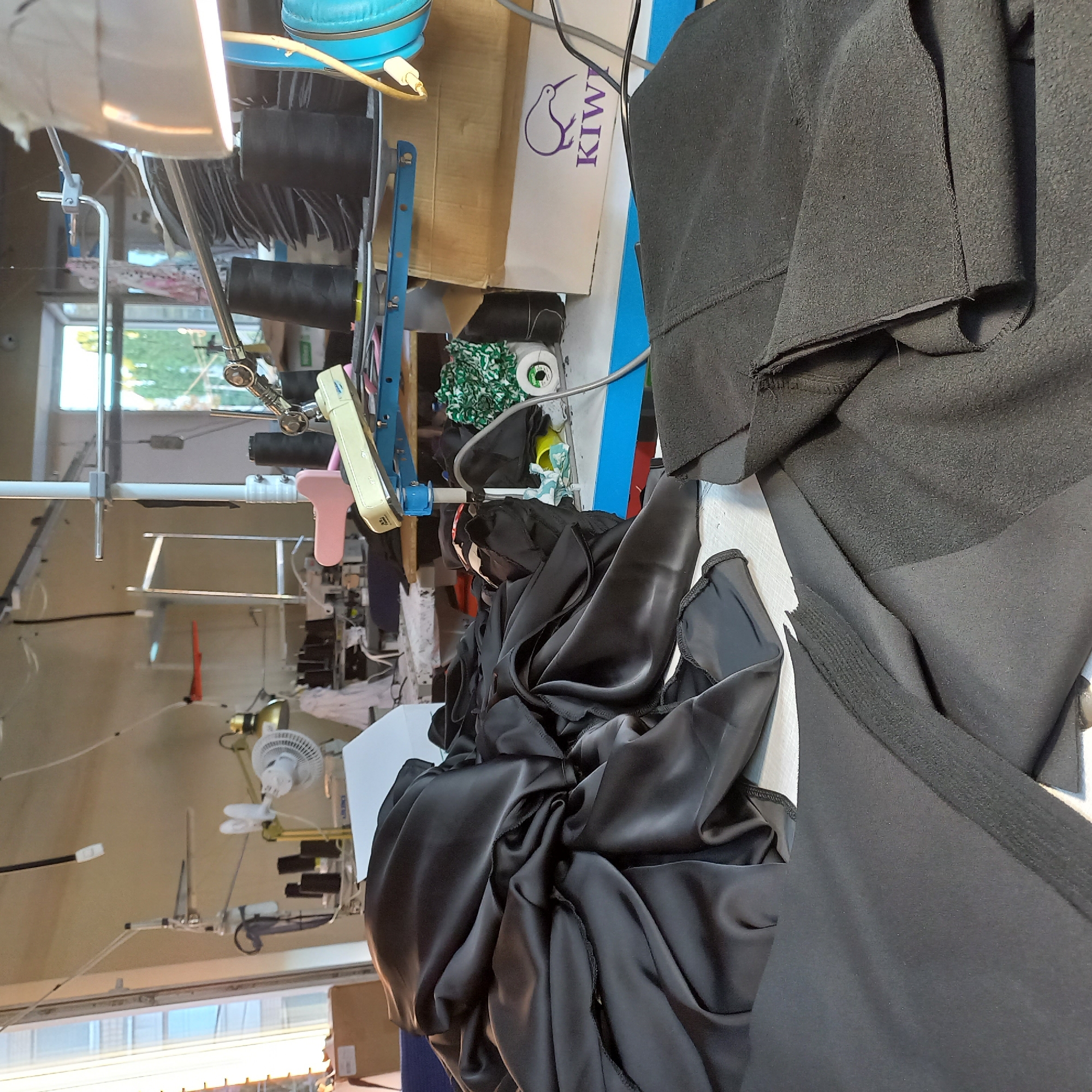 The image depicts a cluttered sewing workshop, likely in Kropyvnytskyi, Ukraine, given the context. A large pile of black fabric, appearing to be satin or a similar slippery material, dominates the foreground. To the right, more black fabric, possibly fleece or a thick cotton, is folded and draped over a surface.
In the background, a sewing machine and various equipment are visible. There are spools of black thread stacked on a blue metal rack, alongside a pink sewing machine foot and a cream-colored sewing machine attachment. A small white fan is positioned near a desk lamp with a gold-colored shade. Further in the background, more sewing machines and industrial equipment are visible, suggesting an active workspace. A cardboard box with a kiwi bird logo and the text "KIWI" is also present on a blue-topped table.
The overall impression is of a functional, albeit somewhat disorganized, sewing studio. The lighting is a mix of ambient light coming from a window on the left and artificial light from overhead lamps. The image is taken from a slightly elevated perspective, looking down onto the work surfaces. There are no people visible in the frame.