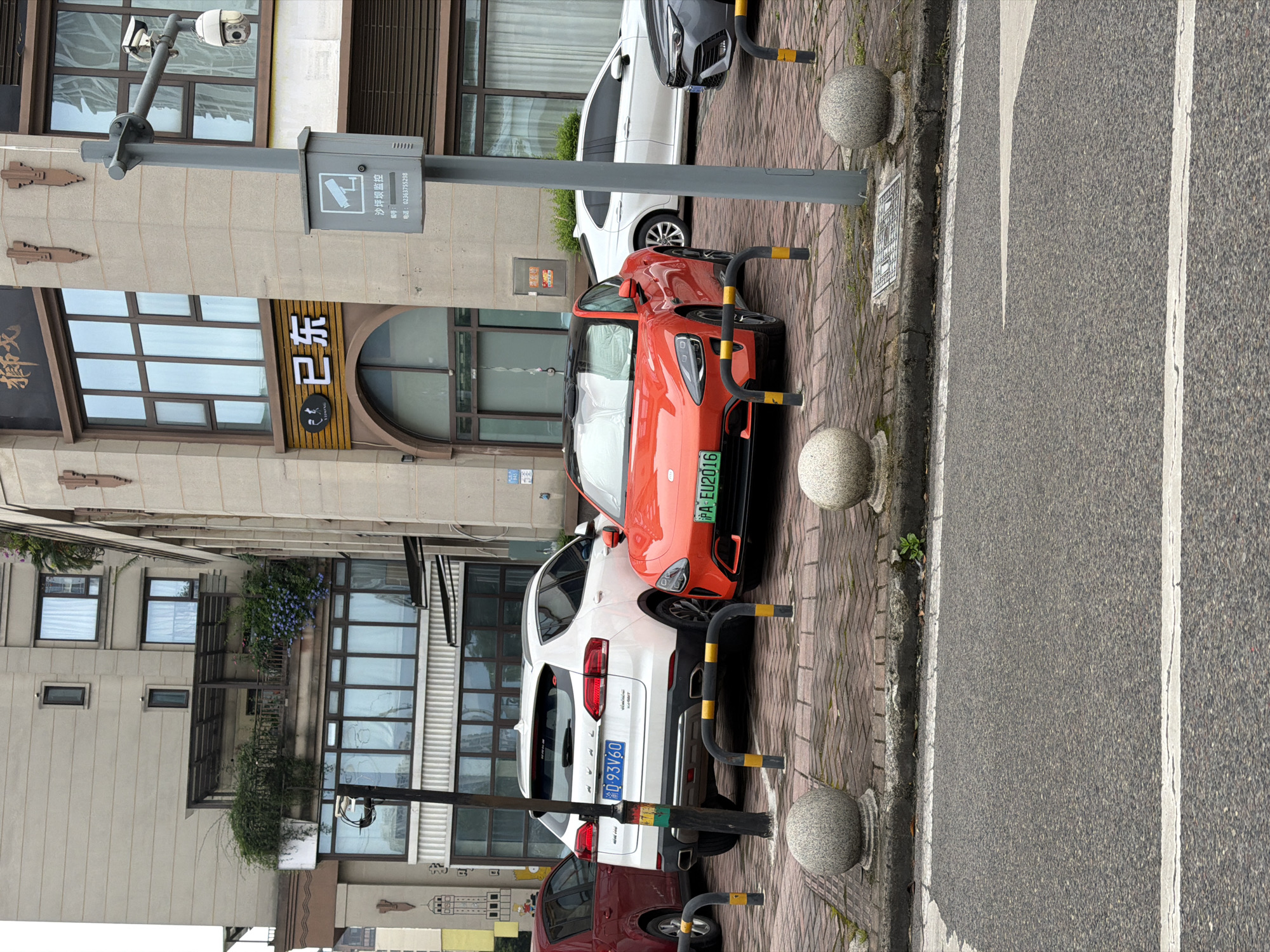 The image depicts a street scene in the High-tech Zone of Chongqing, China. Several cars are parked along the sidewalk, marked by bollards. From foreground to background, the cars are maroon, white, and orange. License plates are visible on the white and orange vehicles; the white car's license plate reads "渝D-93V60" and the orange car's license plate reads "沪A EU2016". The road is to the right, marked with a white line. The buildings are a light-colored stone and have windows and exterior decor such as plants and signage in Chinese characters. There's a surveillance camera attached to a pole. The sign on the pole reads "沙坪坝监控" which translates to "Shapingba Monitoring" and has a phone number: 02363755298.