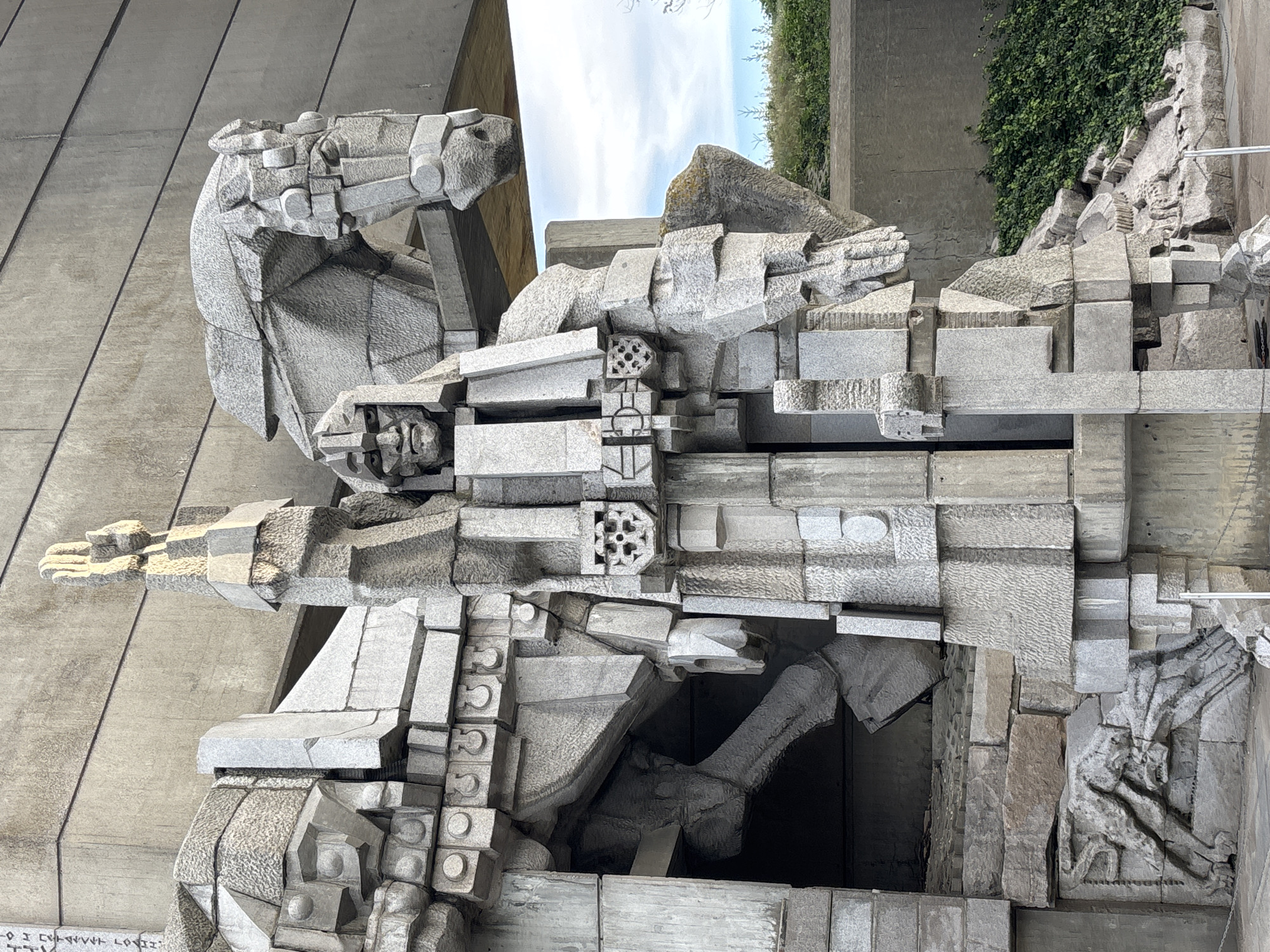 This is a close-up, eye-level, medium shot of a monumental abstract stone sculpture depicting a warrior on horseback. The sculpture is made of rough-hewn, angular blocks of light grey granite. The warrior figure is in the foreground, facing slightly to the left, with its right arm raised and hand open, fingers splayed upwards. The warrior has a stern, stylized face with deep-set eyes and a pronounced brow. Its body is made of geometric blocks, and it wears a segmented helmet and armor. A shield-like emblem with a floral or star pattern is visible on the warrior's chest. Behind the warrior, the head and neck of a horse are visible, also rendered in abstract, angular forms. The horse's eye is a simple circular indentation. The sculpture is massive, with some parts appearing to be carved from the natural rock face. In the lower left of the frame, a bas-relief carving of a rearing horse is visible. The background consists of a textured concrete structure with horizontal lines, and a patch of green foliage and a cloudy sky. The lighting suggests it is daytime, with sunlight casting shadows on the sculpture.