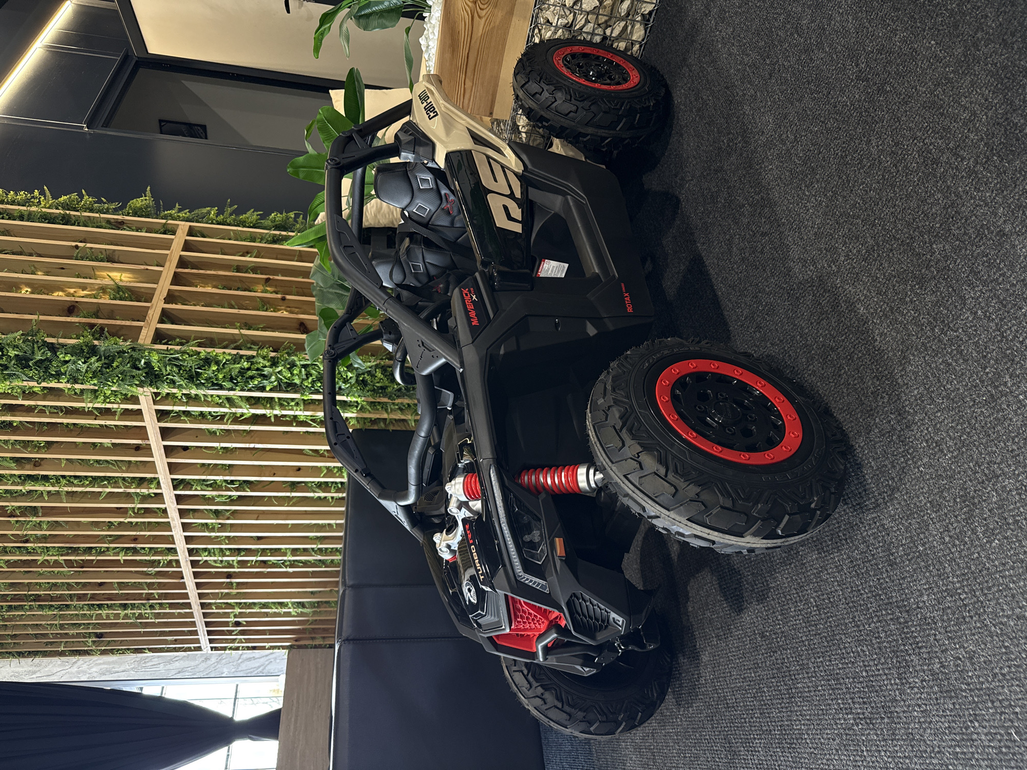 The image shows a black and beige Can-Am Maverick X3 X rs Turbo RR vehicle parked indoors on a gray carpet. In the background, there is a wooden latticework covered with green plants, suggesting a decorative wall feature. A white stone wall is partially visible in the upper right corner. The scene appears to be indoors, possibly in a showroom or exhibit. There is no evidence to suggest a location in Plovdiv, Bulgaria, or any indication of human presence or action.
