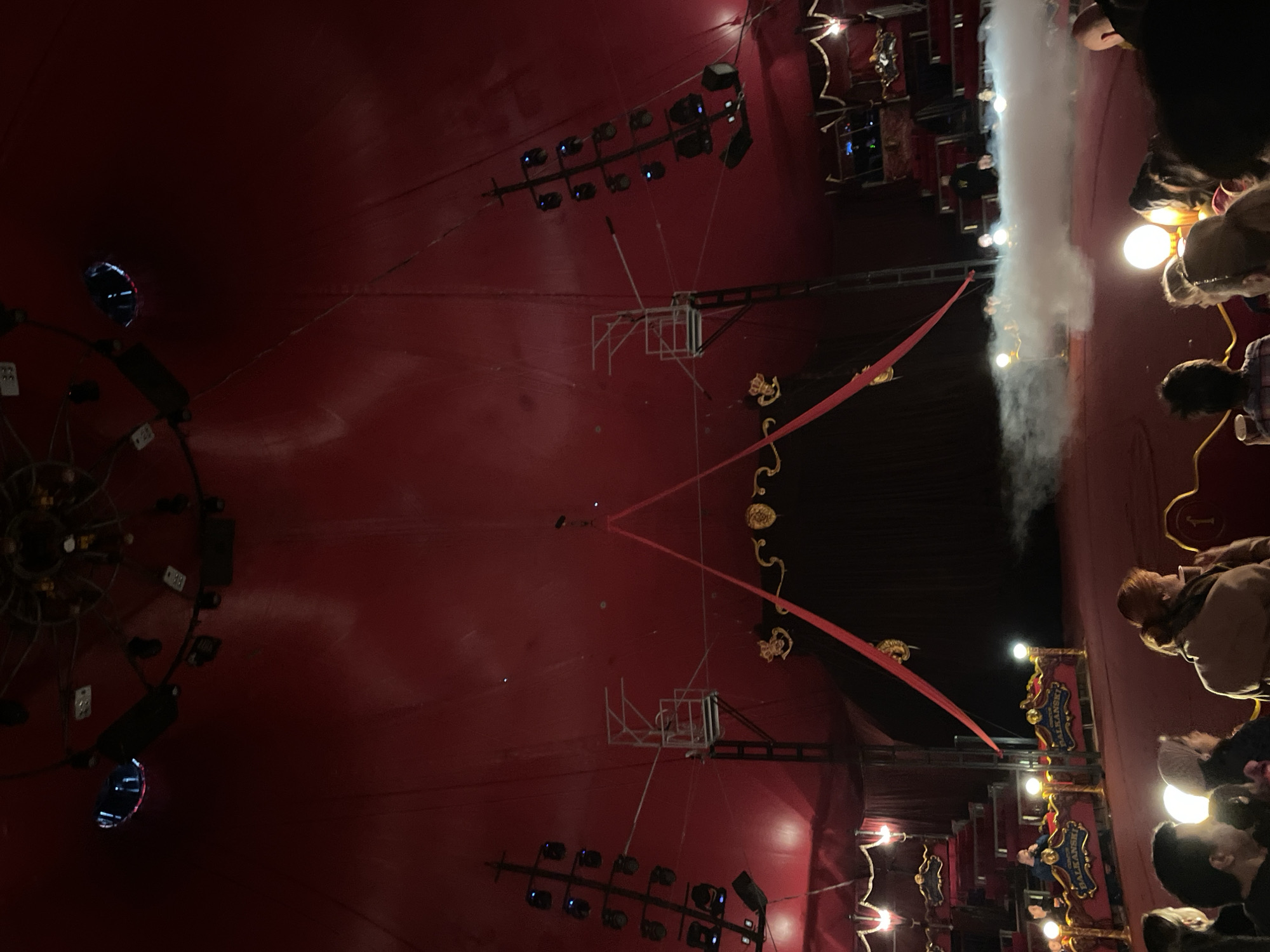 The image shows the interior of what appears to be a circus tent, characterized by a large, red tent ceiling. Lighting fixtures and scaffolding are visible suspended from the tent's structure. Performers and spectators are present, partially obscured by lighting and smoke or haze. A sign bearing the name "Balkanski" is visible. The city of Sofia, Bulgaria, is implied by the tent being a circus.