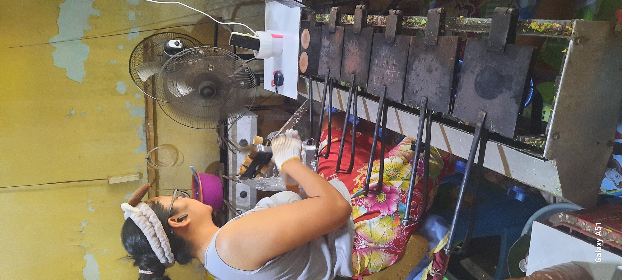 The image depicts a woman, wearing glasses and a headband, seated and engaged in food preparation. She is wearing a grey tank top and a colorful skirt with a floral and butterfly pattern. Her hands are covered with white and yellow gloves, and she appears to be working with a set of long-handled tools aligned over a cooking apparatus. This apparatus consists of multiple dark, square plates with handles, suggesting a specialized cooking method for a specific type of food. Some of the plates are open, revealing what appears to be batter or dough being cooked, while others are closed. A blue flame is visible under one of the plates, indicating it is actively being heated.
The environment surrounding the woman is cluttered and appears to be an outdoor or semi-outdoor workspace. In the background, a large black oscillating fan is prominently displayed, suggesting a warm climate. The wall behind the fan is painted yellow with peeling sections, revealing lighter patches underneath. A pink bowl and various other cooking utensils are visible on a shelf or counter behind the woman. To the right of the fan, a white platform holds a mobile phone in a cylindrical stand and a pair of dark earbuds. The cooking apparatus itself is a metal frame with multiple cooking stations, designed to accommodate several items simultaneously. The overall impression is of a busy, functional food stall or small kitchen, likely operating in a warm climate. The image includes the text "Galaxy A51" in the bottom left corner, indicating the device used for capturing the photograph. The location is identified as Jatitengah, Indonesia.