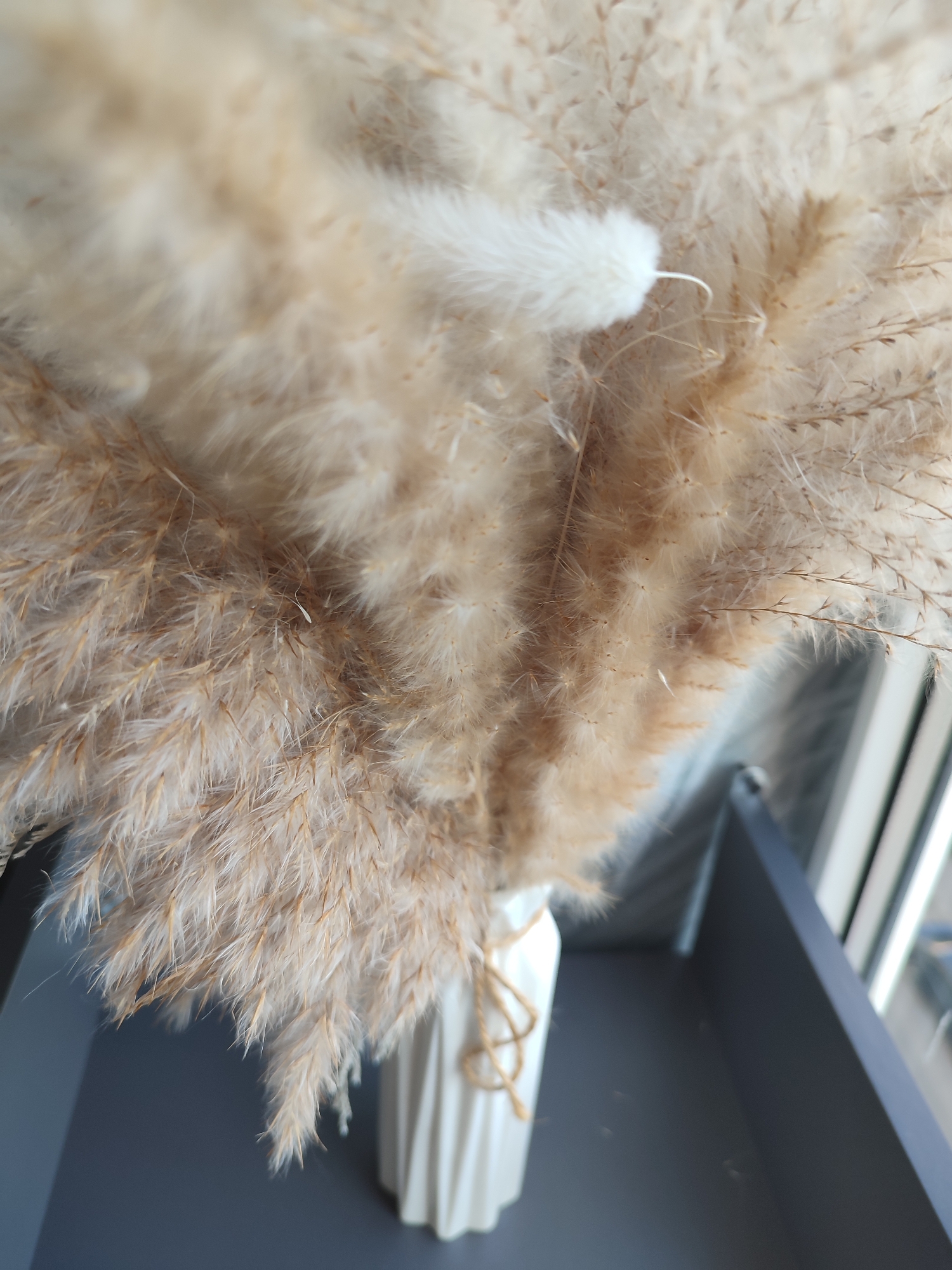 This is a close-up, slightly angled, indoor shot of a dried pampas grass arrangement in a white, fluted vase. The arrangement is predominantly beige and cream-colored, with delicate feathery plumes. A single white feather is prominent among the dried grass. The vase is tied with a jute string. The background shows a dark gray surface, possibly a shelf or table, and a blurry window frame with vertical white and light gray lines, suggesting a window with blinds or panes. The lighting is soft and natural, indicating daylight. There is no discernible location in Voronezh, Russia, based on the provided image.