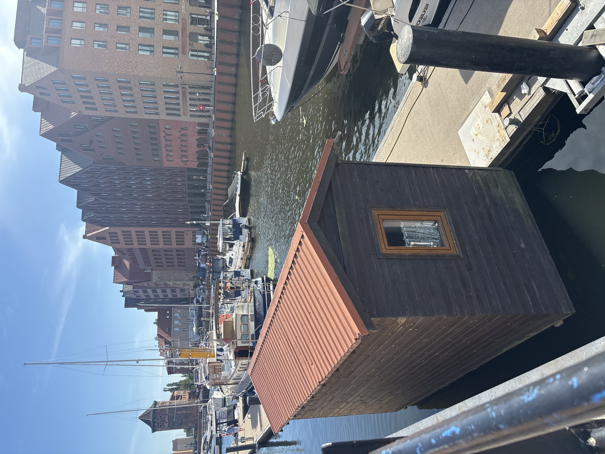 This is a vertically oriented, full-shot photograph taken on a sunny day, likely in the morning or afternoon, given the bright sunlight and clear sky. The scene is a harbor or waterfront with several boats docked and moored. In the foreground, a dark wooden structure with a reddish, corrugated roof sits close to the water. A small window with a lace curtain is visible on the side of this structure.
Further into the harbor, a large white yacht with black trim is prominently docked. Behind the yacht, a series of buildings line the waterfront, appearing to be residential or commercial, with numerous windows and brick facades. The architecture suggests an urban setting. Many other smaller boats, including sailboats and motorboats, are visible in the water, some with their masts reaching high into the sky.
The water appears calm, reflecting the sunlight and the surrounding structures. The overall atmosphere is one of a peaceful, bustling harbor on a pleasant day. No people are clearly discernible in the image, though there may be some figures on the boats or along the waterfront that are too small to identify. There is some faint text visible on the side of the large yacht, but it is not legible.