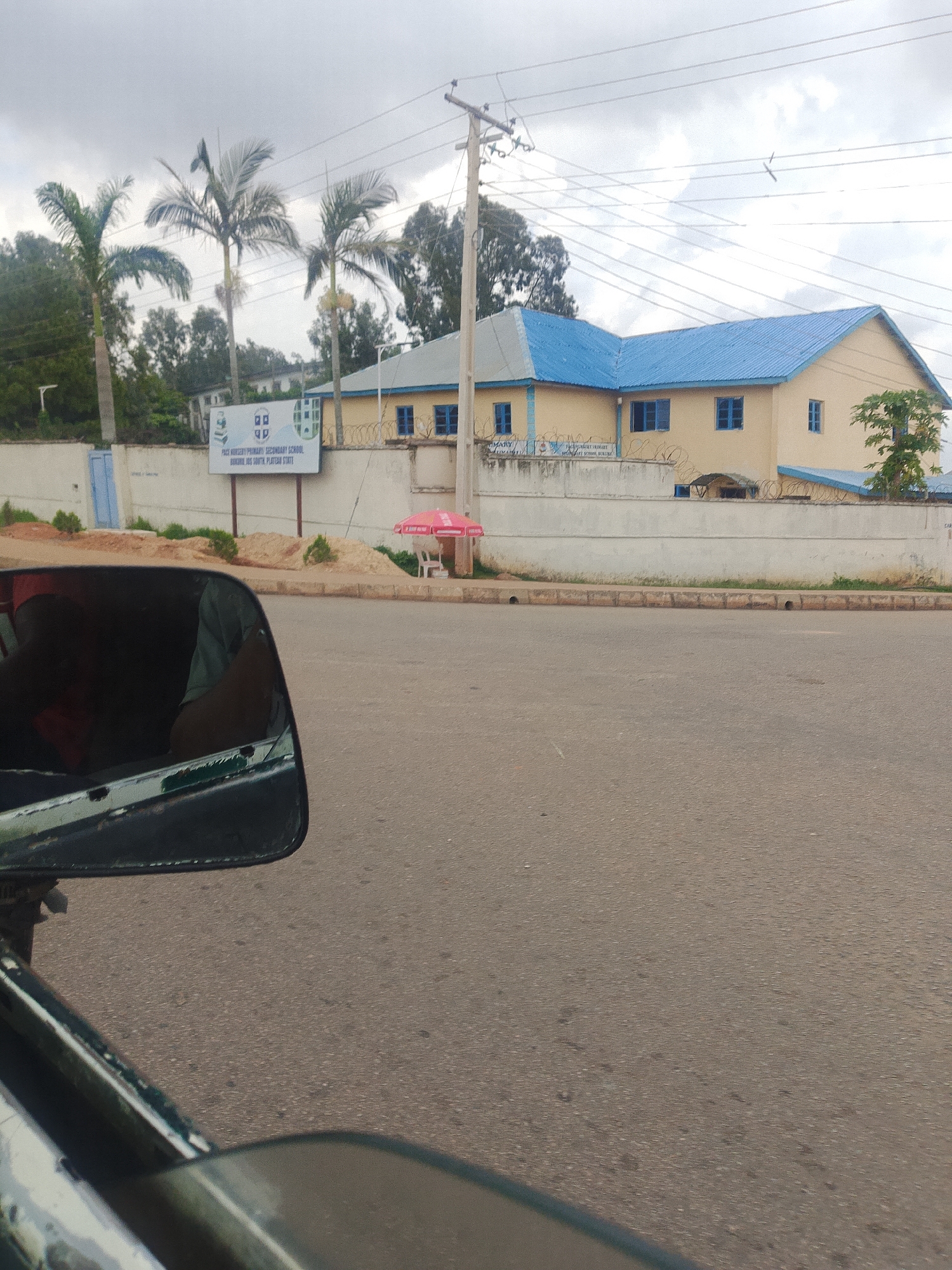 An eye-level, medium shot captures a view from a vehicle, looking out past the side mirror towards a building complex and a street. The building has a light yellow facade and a prominent blue roof. In front of the building, a white wall is visible, topped with barbed wire fencing. A sign on the wall reads "PACK NURSERY PRIMARY SECONDARY SCHOOL BOKKOS JOS SOUTH, PLATEAU STATE". Three palm trees stand to the left of the building. The street in the foreground is paved and appears to be a public road. The sky is overcast.