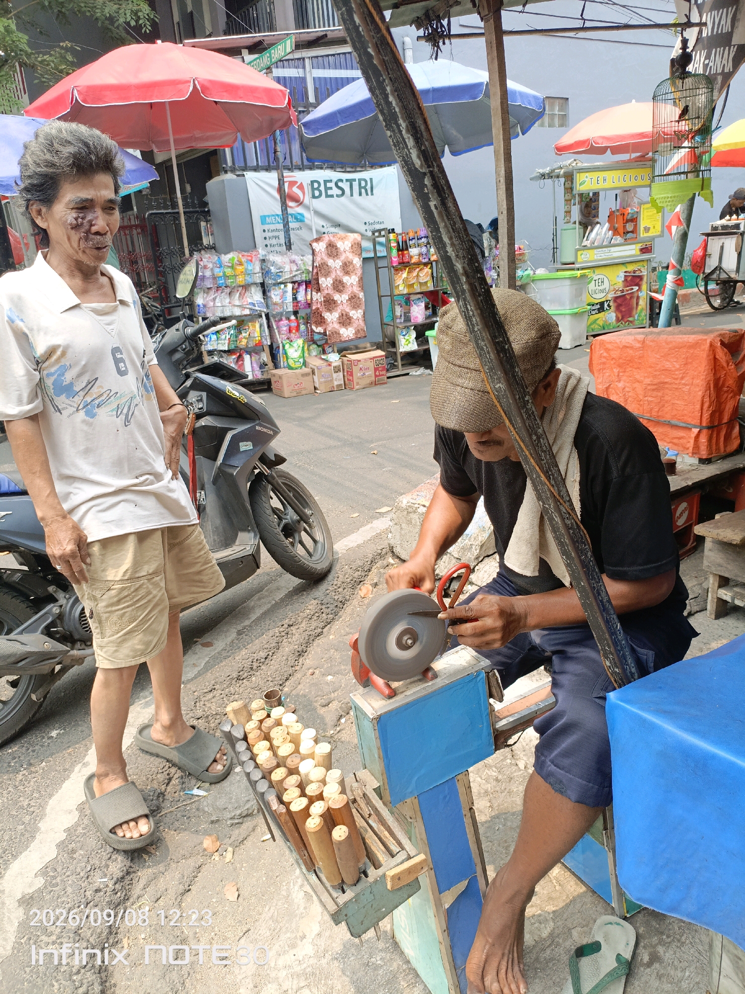 This image captures a street scene in Central Jakarta, Indonesia, likely on a sunny day, given the bright lighting and shadows. In the foreground, a man with weathered skin and grey hair, exhibiting a visible facial injury on the left side of his face, stands next to a motorcycle. He is wearing a light-colored polo shirt with a blue design and khaki shorts, along with grey slip-on sandals. To his right, another man, wearing a flat cap, a black t-shirt with a white towel draped around his neck, and blue pants, is actively sharpening a knife using a grinding wheel mounted on a blue stand. In front of him, a wooden rack holds a collection of wooden-handled tools, possibly for sale. Behind the men, a variety of market stalls are visible, some under red and blue umbrellas, displaying goods such as bottled drinks, cleaning supplies, and patterned fabrics. A birdcage with a bird inside hangs from a pole. The ground is paved with asphalt, with some scattered leaves. A watermarked date and time stamp at the bottom of the image reads "2026/09/08 12:23" and "Infinix NOTE30".