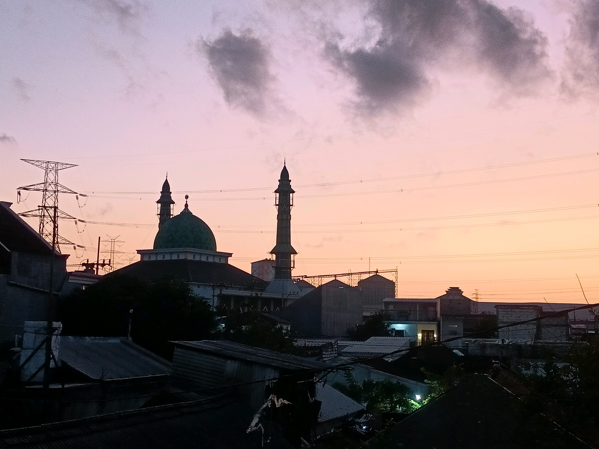This photograph captures a twilight scene over the city of Ngargosari, Indonesia, with a focus on a mosque and surrounding urban landscape. The sky displays a gradient of soft pinks and purples, transitioning to a warm orange near the horizon, indicative of sunset or sunrise. Prominent in the mid-ground is a mosque with a distinct green dome and a tall, slender minaret, silhouetted against the colorful sky. To the left, a large electricity pylon stands tall, its metal lattice structure starkly contrasting with the softer hues of the sky. The foreground is dominated by the dark silhouettes of rooftops of residential or commercial buildings, their corrugated metal and tiled surfaces creating a textured layer. In the distance, industrial structures and other buildings are visible, suggesting a mixed-use urban environment. Power lines crisscross the frame, adding to the urban feel. No people are discernible in the image, and the scene is static, conveying a sense of quietude typical of dusk in a populated area. The overall impression is one of urban development juxtaposed with a prominent religious structure under a dramatic sky.