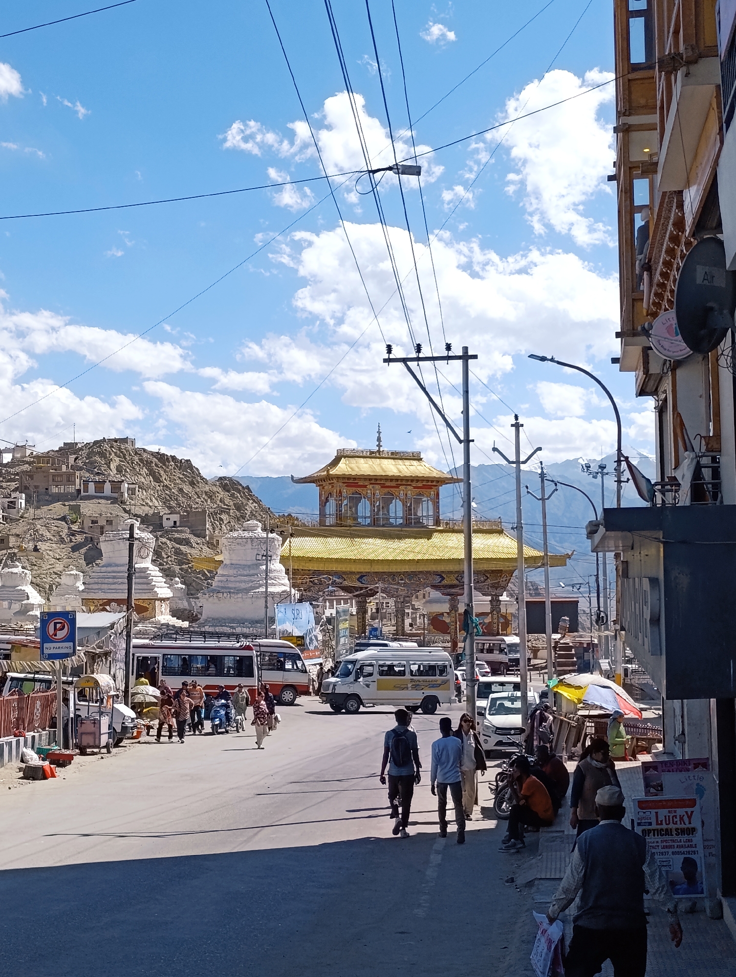 This is a ground-level, eye-level shot of a street scene in Leh, India, likely in the vicinity of the main market area, near the Tsemo Fort or Leh Palace. The image captures a bustling street with a mix of vehicles and pedestrians under a bright blue sky dotted with fluffy white clouds.
In the foreground, the asphalt road is marked with shadows. Several people are walking on the right side of the street, some on the sidewalk. One person in the immediate foreground is walking away from the camera, carrying bags. Further up the street, on the right, a shop is visible with a sign that reads "LUCKY OPTICAL SHOP".
On the left side of the street, a bus and several other vehicles are parked or moving. A "No Parking" sign is visible. Beyond the vehicles, several white stupas with layered roofs are prominent, with a complex of buildings perched on a rocky hill in the background, indicative of a traditional Leh architectural style.
Dominating the mid-ground is a large, ornate gateway structure with a golden roof, characteristic of Buddhist architectural styles found in the region. This structure appears to be a significant landmark. Power lines crisscross the sky, a common sight in urban environments. The overall impression is one of a vibrant, culturally rich location in a mountainous region.