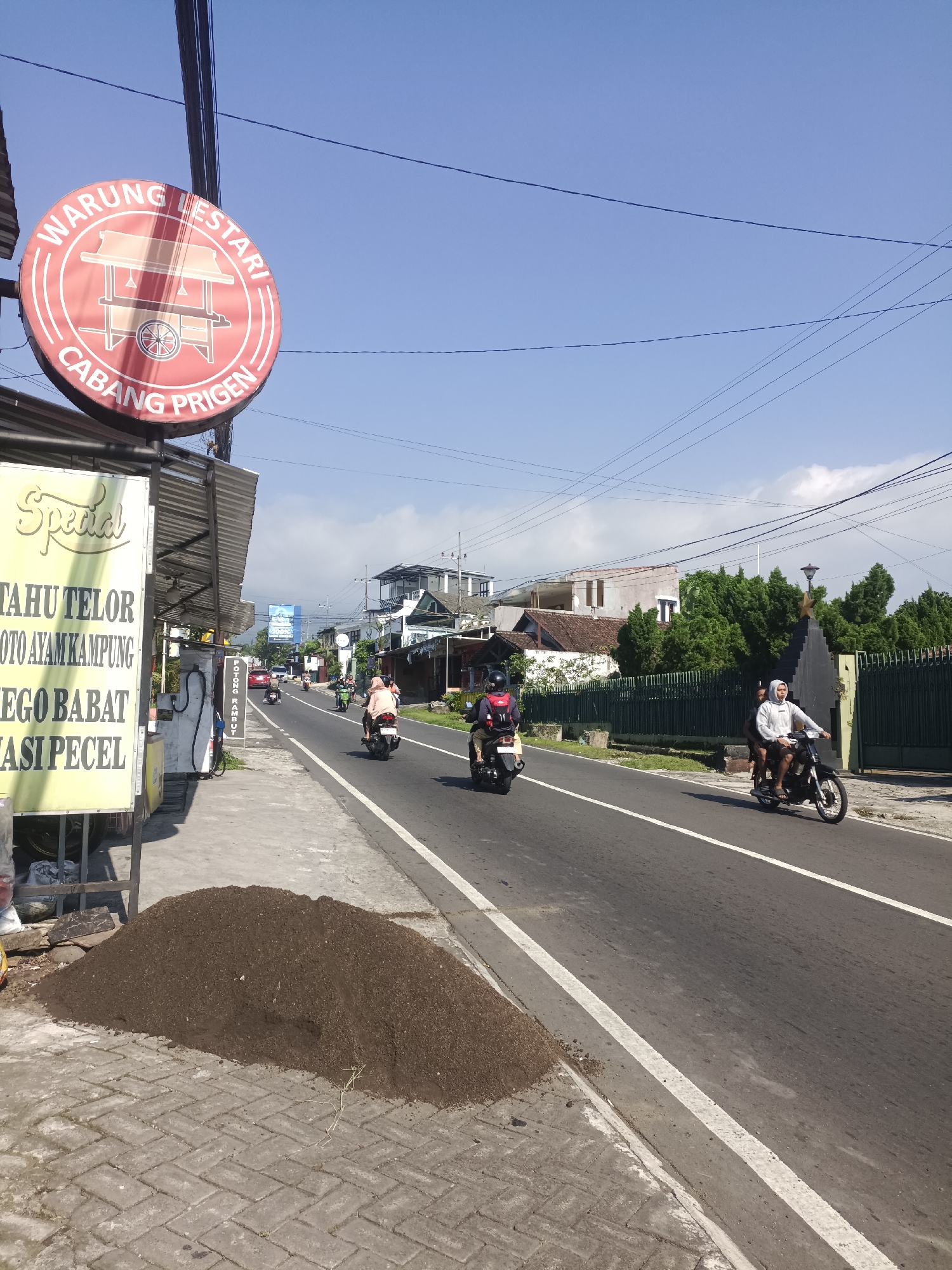 The image depicts a street scene in Prigen, Indonesia, under a clear sky. A "Warung Lestari Cabang Prigen" sign is visible on the left. Below the sign is a menu advertising "Special Tahu Telor, Oto Ayam Kampung, Ego Babat, and Nasi Pecel". A pile of dirt or soil sits on the sidewalk near the menu board. Motorbikes travel along the road, each carrying one to three passengers. Buildings and greenery line the roadside. Overhead, power lines stretch across the sky.