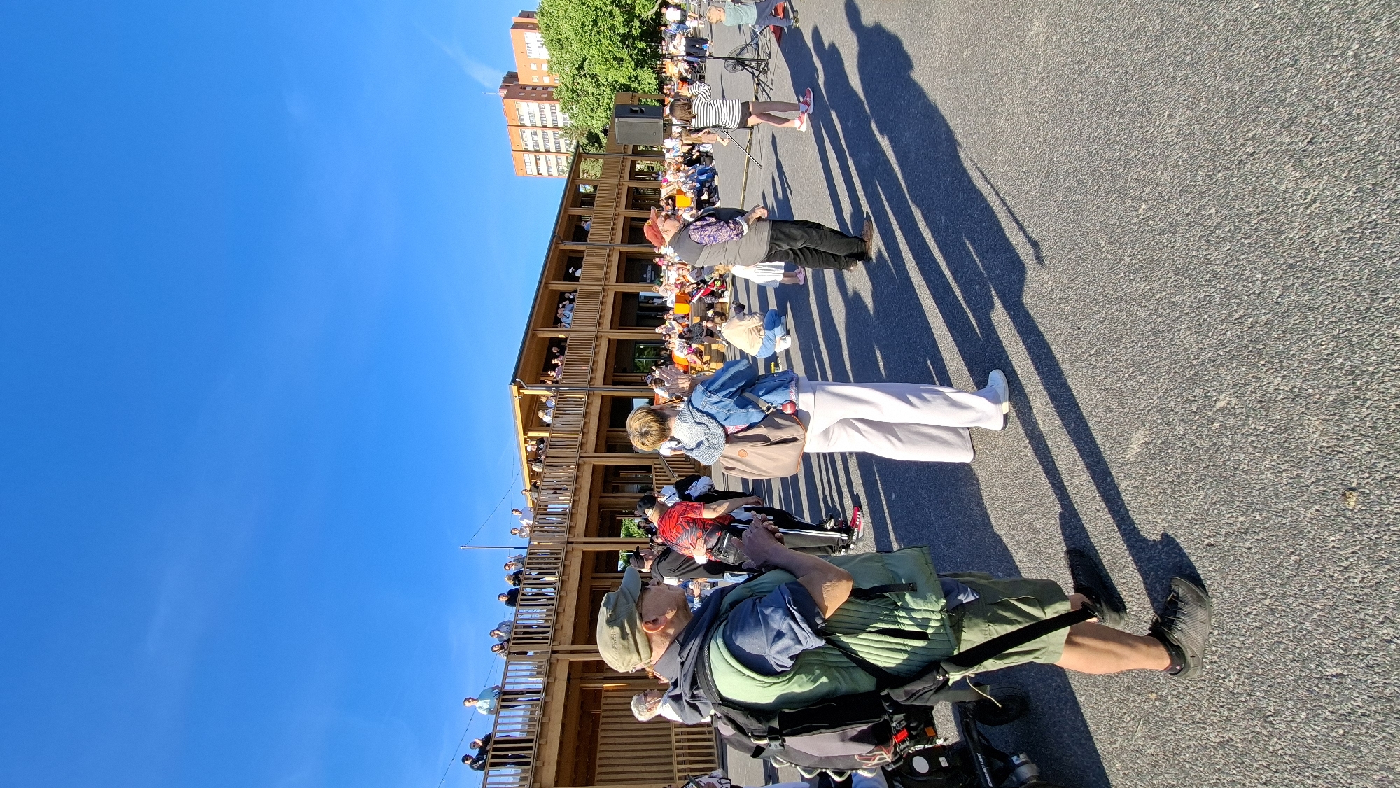 The image shows an outdoor gathering with a distinct wooden structure resembling a tiered stage or seating area. Many people are present, some seated on the tiers of the structure and others standing or sitting on the asphalt ground. The sky is a clear, bright blue, suggesting it is daytime, likely sunny given the long shadows cast on the ground, indicating either morning or late afternoon.
In the foreground, a person in a wheelchair is visible, wearing a green vest and a baseball cap. Several other individuals are also in the foreground, some standing and looking towards the wooden structure, while others are seated. The wooden structure itself is multi-leveled and appears to be a focal point of the gathering, with people observing something or someone from its various levels.
Further back, a woman in a striped dress stands in front of a microphone and speaker, suggesting a performance or presentation is taking place. A crowd is gathered around her, many appearing to be attentive. In the background, a multi-story brick building and trees are visible, providing some context to the surrounding environment. The overall atmosphere seems to be one of a public event or festival. No visible text is present in the image.
