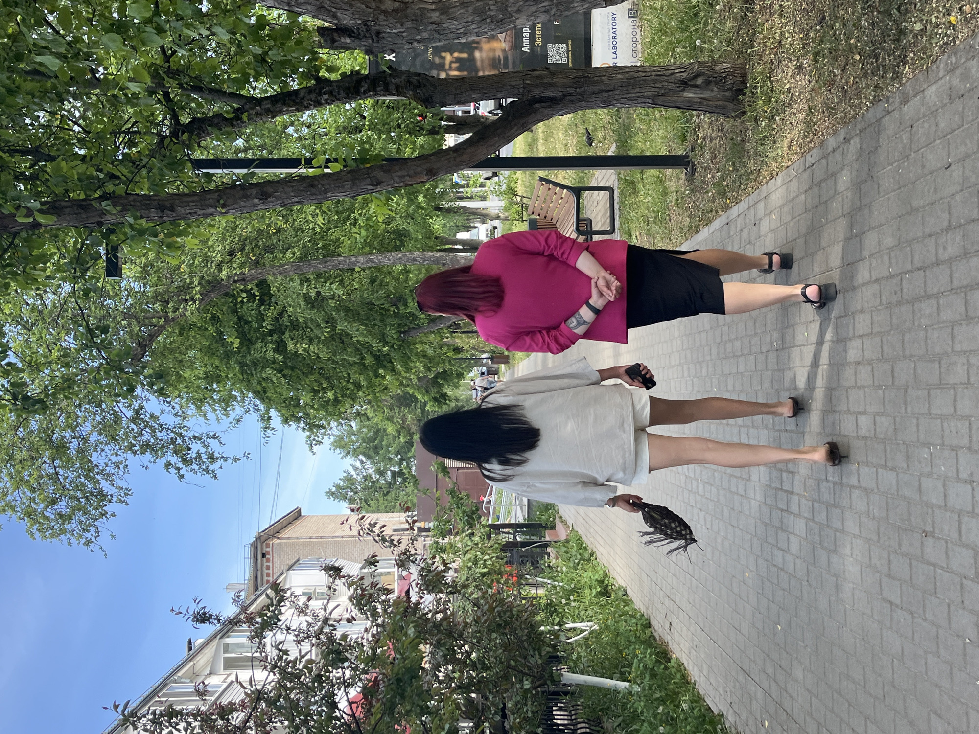 Two women walk side-by-side on a brick sidewalk in Chaikovsky, Russia. The first woman, closer to the foreground, has long, dark hair, wears a white top and shorts, and carries a dark object in her left hand. She also has a phone in her right hand. The second woman, slightly behind, has auburn hair, wears a pink top and a black skirt, and has her hands clasped behind her back. Both women wear sandals. A bench is visible on the right side of the sidewalk, with trees providing shade. Buildings are visible on the left, and a sign can be seen on the right. The sky is partially visible through the trees.