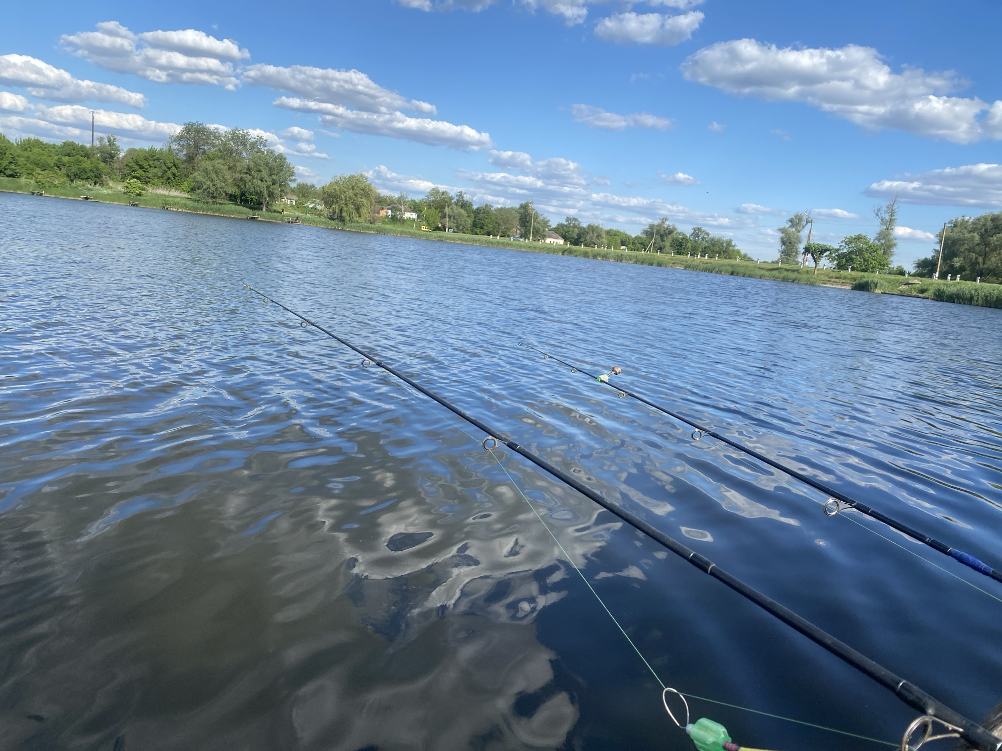 The image depicts a body of water, likely a lake or large pond, with fishing rods extended over the water. The surface of the water is rippled, reflecting the sky and clouds above. The shoreline is visible in the background, with trees and vegetation along the edge. The sky is blue with scattered white clouds. The location is in Ustyvytsia, Ukraine. There are no people visible in the image.