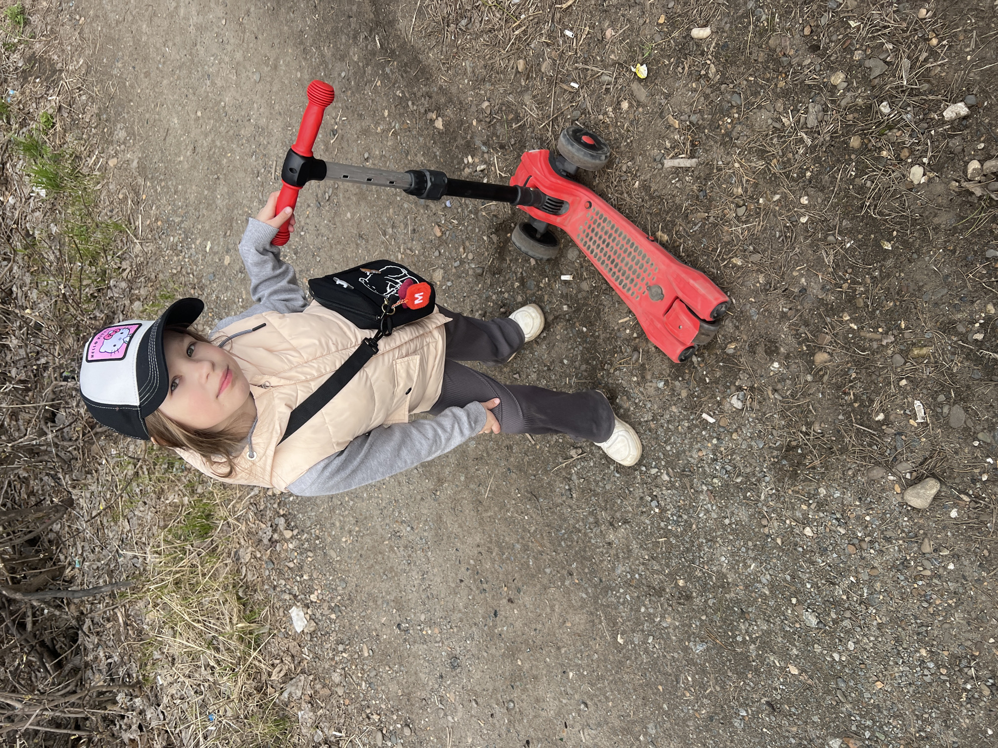 A young child stands on a dirt and gravel path, holding a red three-wheeled scooter. The child looks directly at the camera with a neutral, calm expression. She wears a black and white baseball cap with a "Hello Kitty" patch on the front, a light beige quilted vest over a grey long-sleeved shirt, dark grey pants, and white sneakers. A black shoulder bag with a white graphic, possibly a stylized animal, and a red pom-pom charm with a white 'W' or 'M' is slung across her body.
The setting is an outdoor area in Chaikovsky, Russia, characterized by a well-trodden path composed of loose dirt, small stones, and dry organic matter like twigs and dead grass. Sparse green and dry vegetation lines the sides of the path. The red scooter, equipped with a black adjustable handlebar and black wheels, shows some signs of wear. The lighting suggests daytime, possibly under an overcast sky or with diffused sunlight, as there are no harsh shadows. The image captures a candid moment during an outdoor activity like scootering.