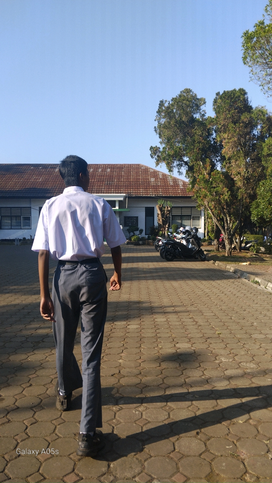 A young male student, viewed from behind, walks away from the camera down a paved walkway. He is wearing a white short-sleeved school shirt and dark grey trousers. The walkway is constructed of hexagonal pavers with a diamond pattern in the center. To the left of the walkway is a low, long building with a reddish-brown tiled roof. The building has multiple windows. To the right of the walkway are several trees with green foliage, and parked motorcycles. The sky is clear and blue. The image is watermarked with "Galaxy A05s" in the bottom left corner. The scene appears to be an outdoor area of a school or similar institution. This photograph was taken in Sumedang, Indonesia.