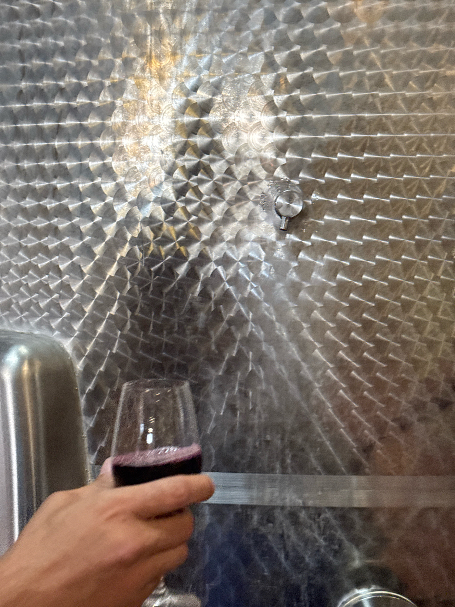 In Kurdghelauri, Georgia, a person's hand is holding a glass of red wine. The glass is positioned in front of a large, textured metal surface, likely a wine fermentation tank. The metal has a repeating diamond pattern that reflects light, creating a shimmering effect. A small valve or spigot is visible on the metal surface. The scene appears to be set in a winery or cellar.