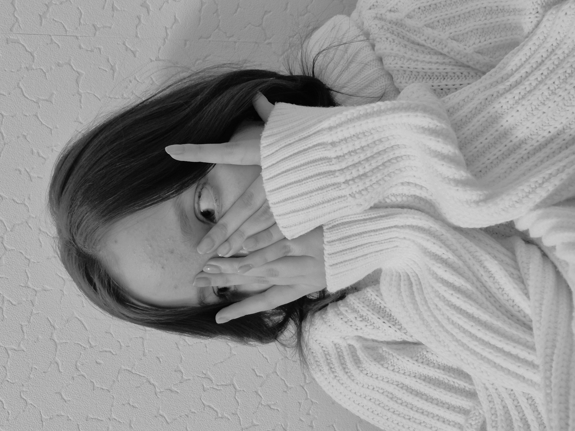 A grayscale, close-up studio shot depicts a person, possibly female, with dark hair, covering part of their face with their hands. The person is wearing a light-colored, heavy knitted sweater. The background appears to be a textured wall. No specific location or additional contextual details pertaining to the city of Chaikovsky, Russia, are evident.