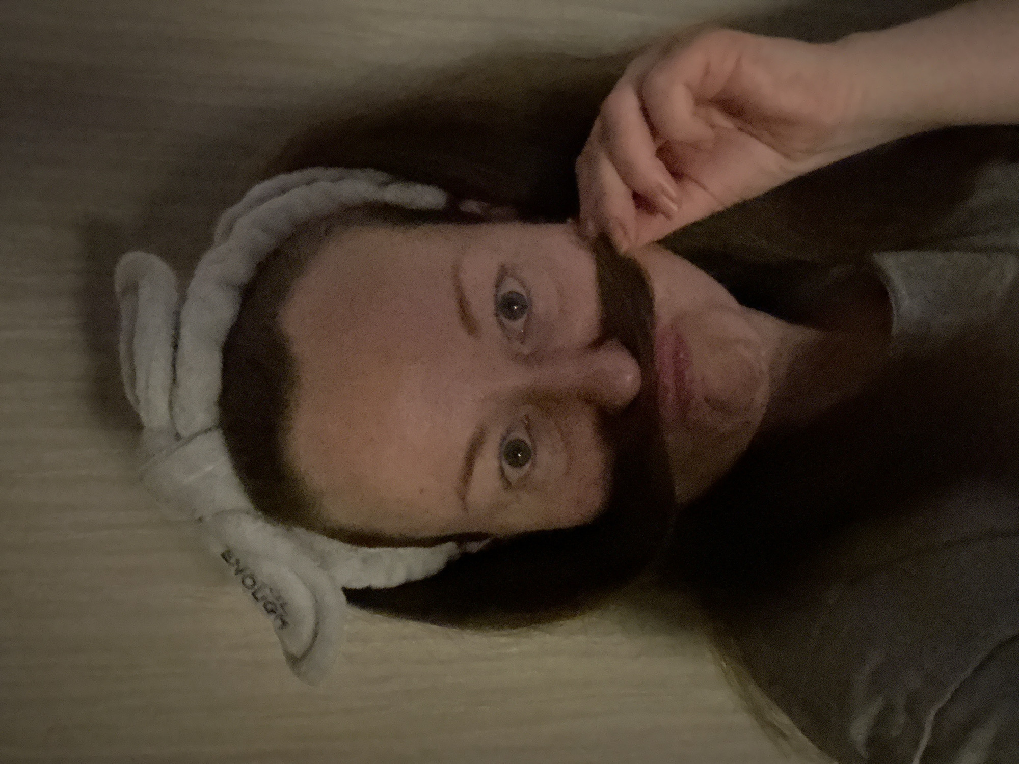 A woman with brown hair is lying on a light brown surface, possibly a wooden floor. She is wearing a light gray headband with the word "ENOUGH" visible. She is holding a strand of her hair to resemble a mustache. She is wearing a dark gray shirt. The location is not discernible from the image.