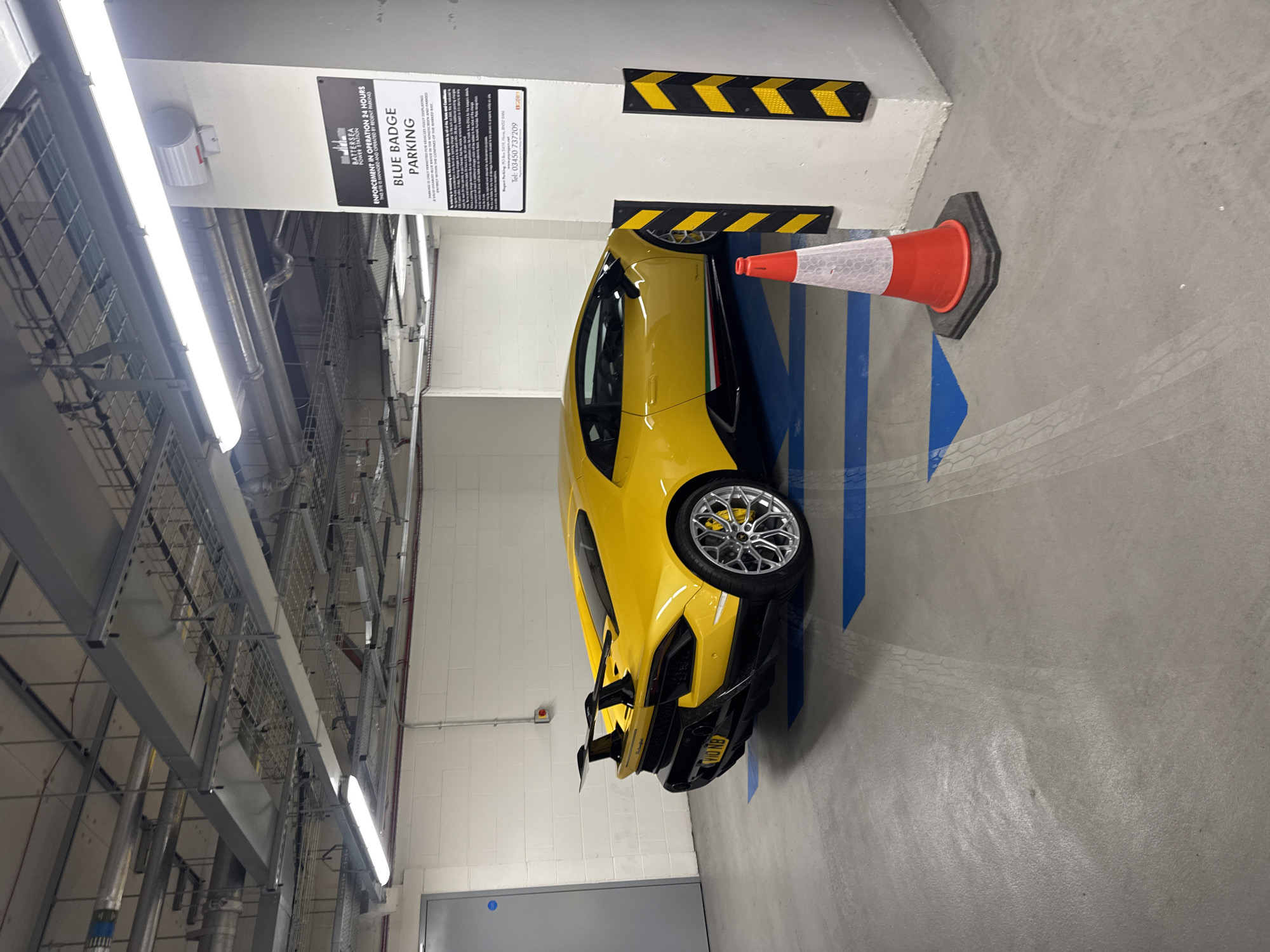 The image shows a yellow sports car parked in a "Blue Badge Parking" spot in what appears to be an underground parking garage at Battersea Power Station in London, United Kingdom. The car is parked next to a wall with black and yellow striped bumper guards and an orange traffic cone. A sign on the wall specifies that parking is for "Blue Badge" holders and the site is managed by Regent Making. The garage has a gray floor with blue painted parking lines. Above, there's an exposed ceiling with a complex network of pipes, cables, and metal supports.