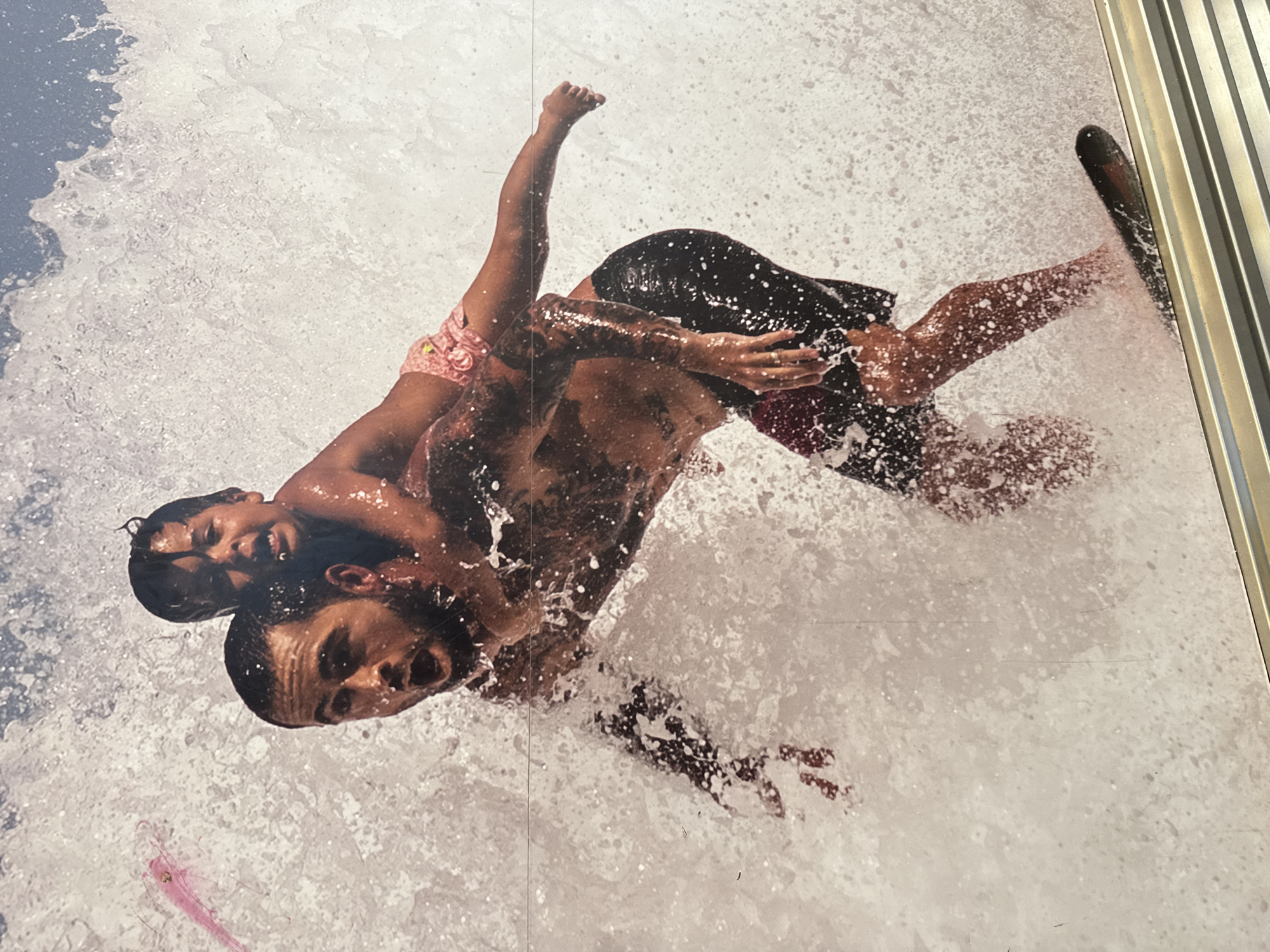 This vibrant image captures a dynamic scene featuring two individuals, an adult male and a child, immersed in a powerful splash of white, turbulent water. The subjects appear to be in mid-action, suggesting an intense water sport like surfing, bodyboarding, or being enveloped by a large wave.
The adult male, sporting dark hair and tattoos on his arms and torso, is clad in dark swimming shorts. He is carrying or supporting the child, who is positioned on his back or shoulders and wears light-colored, patterned swimming shorts. Both figures are visibly wet, with water droplets clinging to their skin. Their facial expressions convey excitement: the child's mouth is open in what seems to be a joyful shout or laugh, while the adult displays a focused and exhilarated look. A sliver of blue, likely sky or calmer water, is visible in the upper left corner.
The media file itself appears to be a photograph of a large-scale print or mural, evident from faint vertical seam lines that traverse the image and a distinct metallic vertical strip framing the right edge, suggesting it is part of an architectural or display installation. The bright lighting implies a sunny day. There is no visible text in the image. This photograph was captured in Buenos Aires, Argentina.