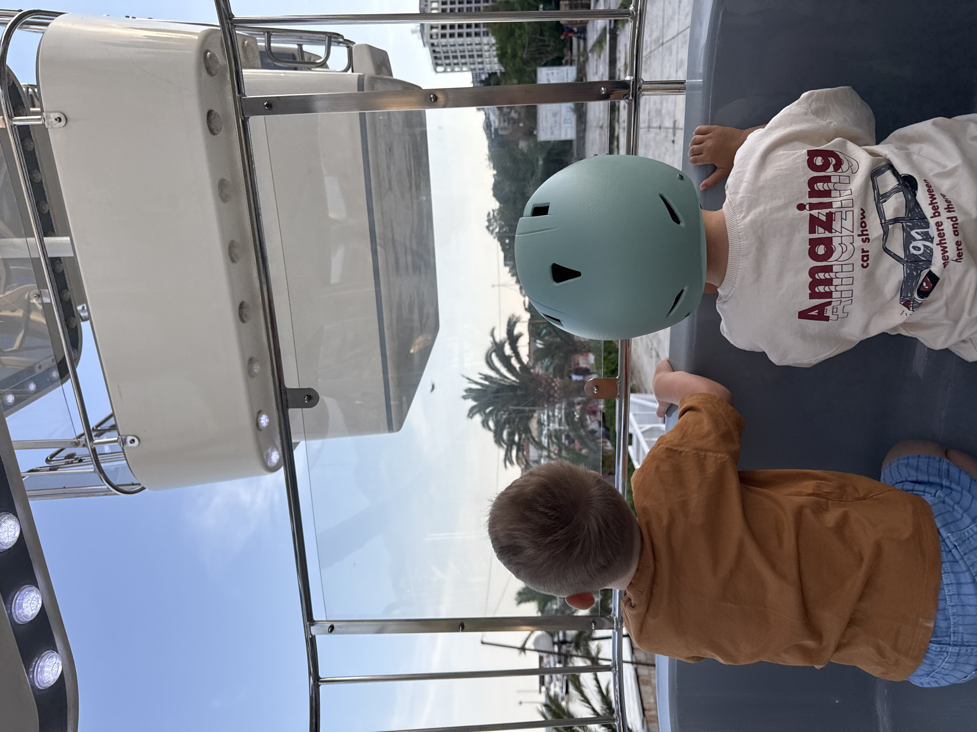 Two young children are observed from behind inside an enclosed cabin or gondola, likely part of an amusement ride such as a Ferris wheel or cable car. The child on the right wears a mint green helmet and a white t-shirt with a graphic design. The t-shirt features the prominent text "Amazing car show" along with a drawing of a classic car and smaller text "91 somewhere between here and there." The child on the left wears a brown long-sleeved shirt and blue plaid shorts. Both children are looking out through the clear glass panels of the enclosure, observing the scenery outside.
The setting is outdoors during the daytime, under a clear blue sky with a few wispy clouds. Through the glass, palm trees are visible, suggesting a warm climate. In the distance, several buildings can be seen, hinting at an urban or resort area. The structure of the enclosure includes metal railings, clear panels, and white solid panels adorned with small lights. The children appear to be engaged in observing the panoramic view from their elevated position. The time of day seems to be late afternoon or early evening due to the quality of light. The overall scene depicts a leisurely moment of observation from an attraction in Budva, Montenegro.