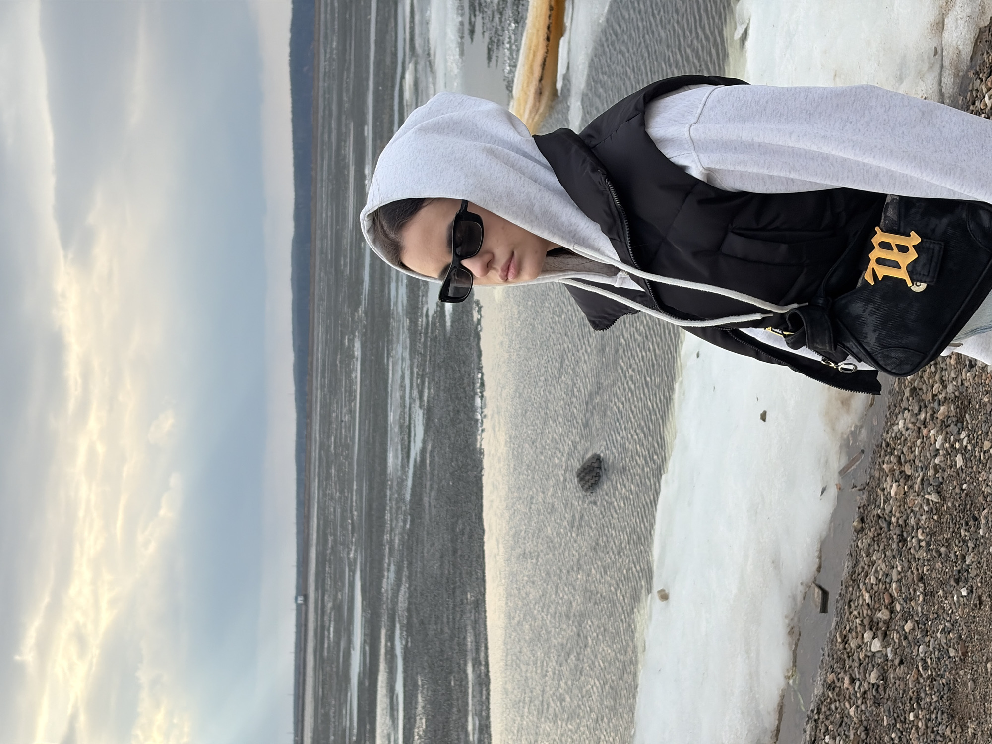 A person in a grey hoodie and black vest stands next to a body of water, likely the Kama River, near Chaikovsky, Russia. The background includes a pale sky and the partially frozen river. The foreground shows a gravelly shoreline. The person wears sunglasses and a black purse with a gold emblem.