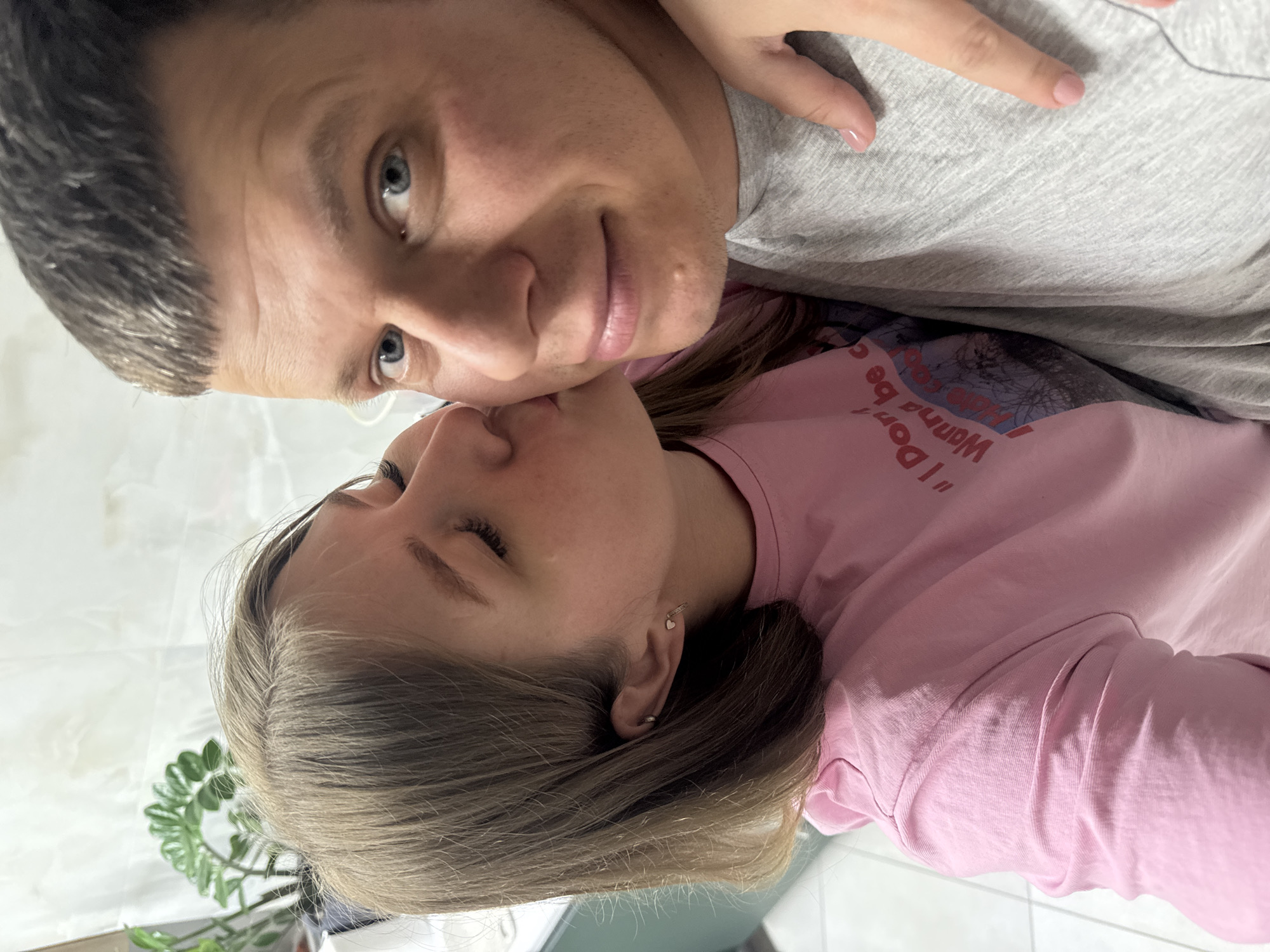 The image shows two people posing closely together. A man with short, dark hair and light eyes is wearing a gray shirt and looking directly at the camera with a slight smile. A woman with long, light brown hair and closed eyes is wearing a pink long-sleeved shirt. They appear to be close, possibly kissing or nuzzling. The background is blurry but suggests an indoor setting, with hints of a plant and a white surface. This scene is located in the city of Gornyi (Dvurechenskoe S/P), Russia.