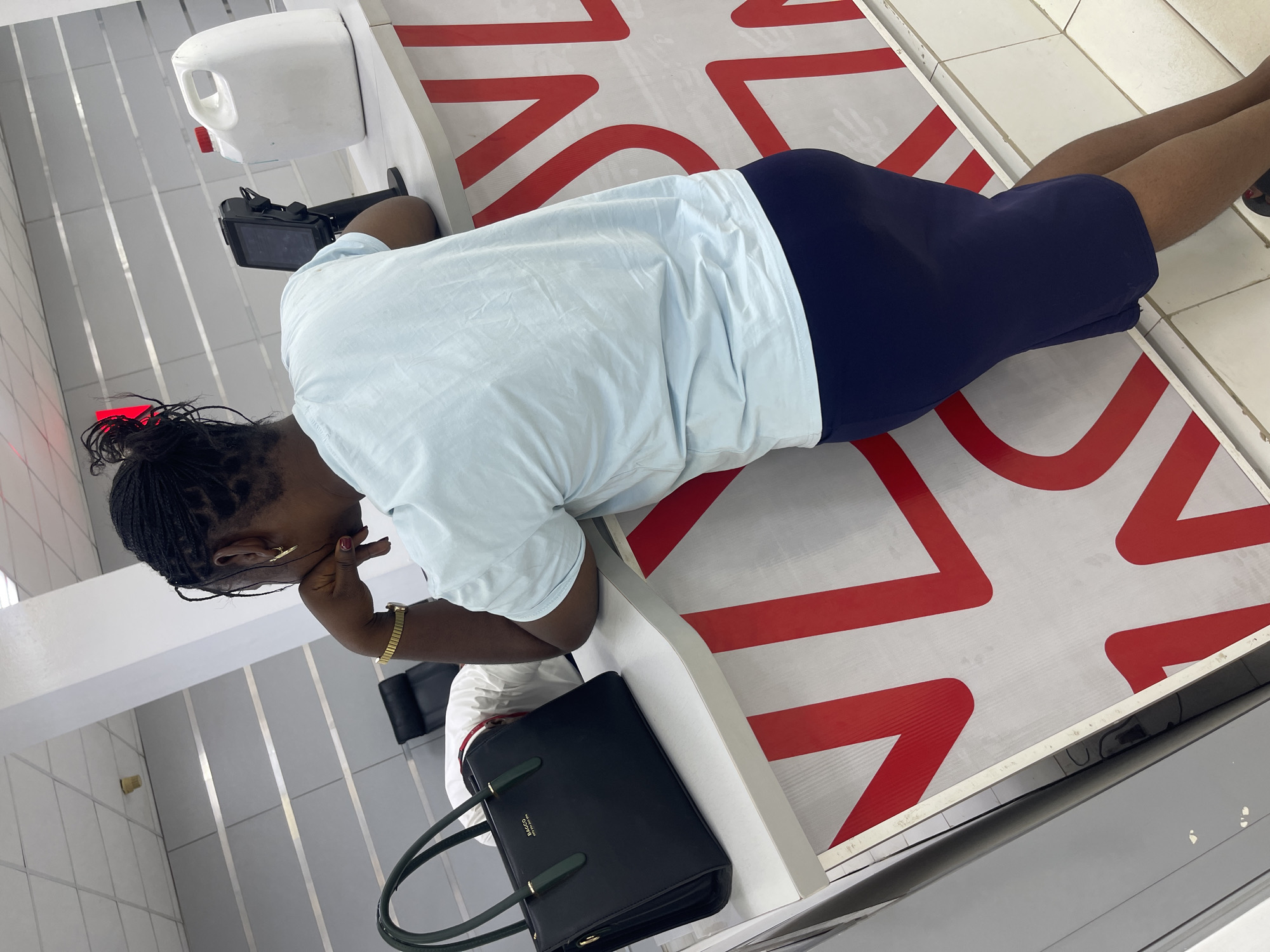 A woman with dark skin and braided hair sits resting her head on her hand at a checkout counter in a store. She wears a light blue t-shirt and a dark blue skirt. On her wrist is a gold wristwatch. Beside her is a black handbag with green handles. The checkout counter is decorated with a red and white pattern. Behind the counter is a white dispenser. The location appears to be in a commercial space, potentially a retail store in Zaria, Nigeria, though this cannot be confirmed with certainty based on the image alone.