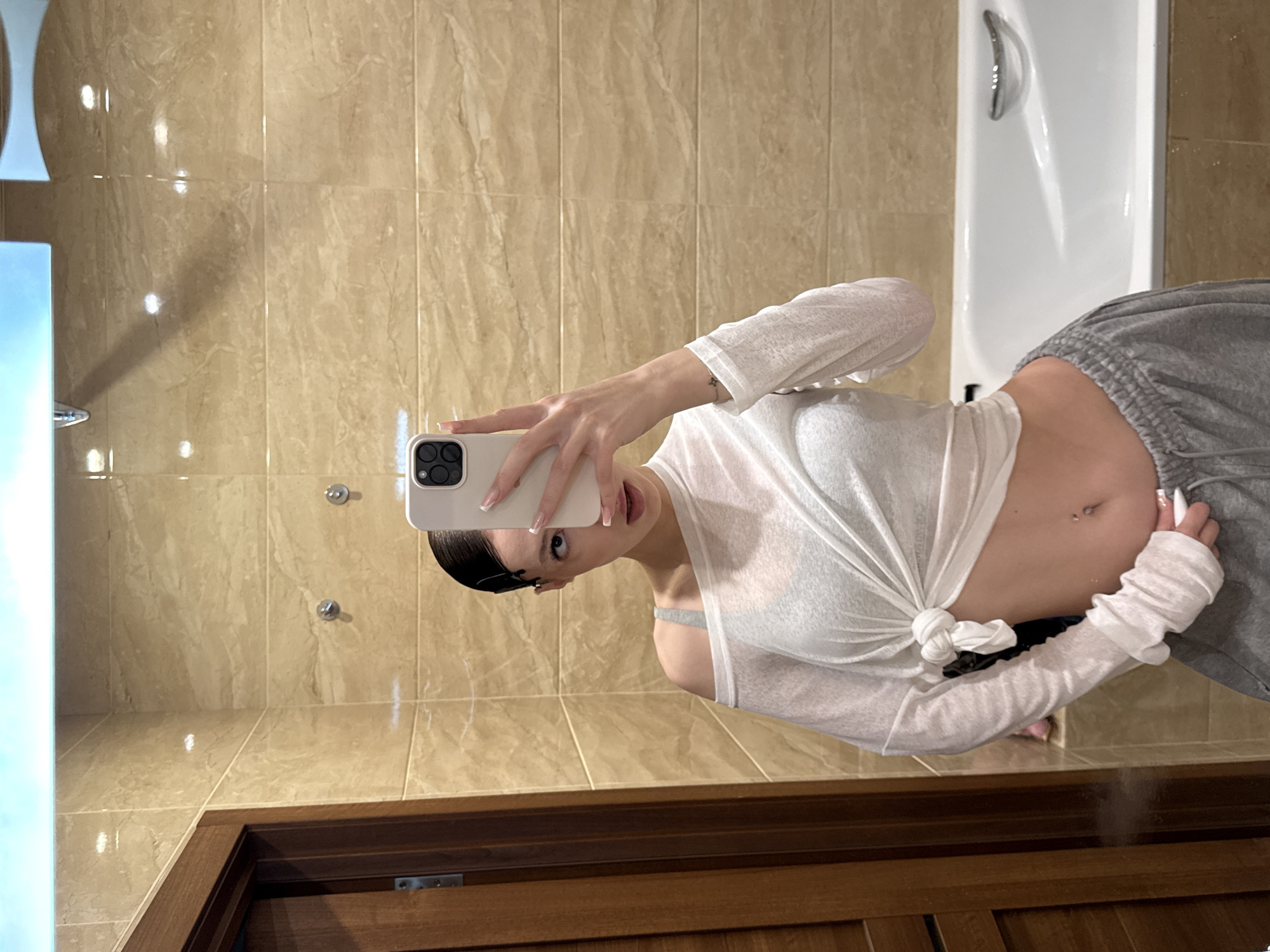 A young woman with dark hair pulled back is taking a mirror selfie in a bathroom. She is wearing a white sheer long-sleeved shirt tied at the waist, revealing a grey bra and her midriff with a belly button piercing. She is also wearing grey sweatpants. The bathroom features beige tiled walls, a wooden door on the left, and a white bathtub with a chrome handle on the right. The lighting suggests an indoor environment, likely a residential bathroom. The image appears to be taken in Khimki, Russia.