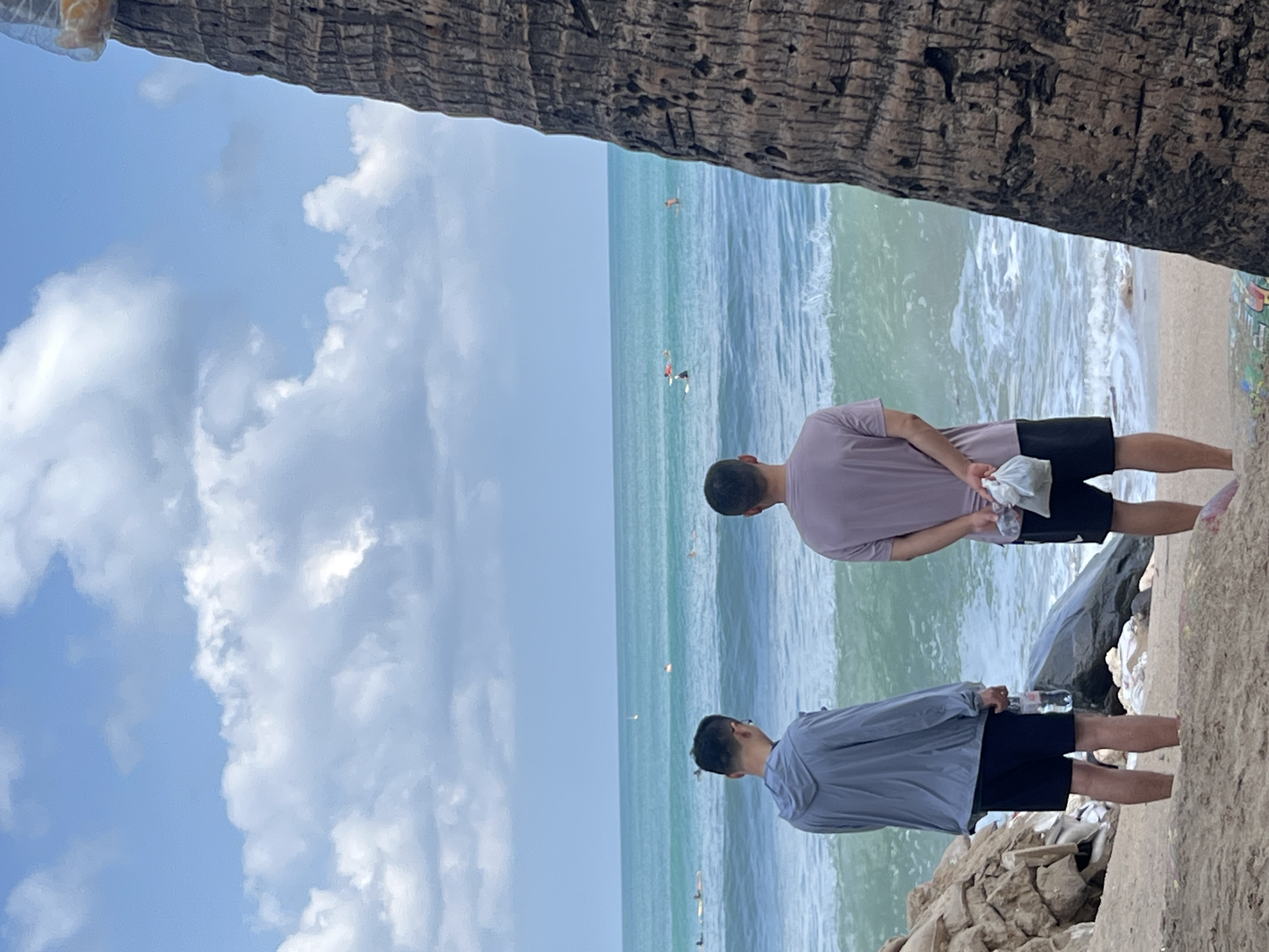 Two individuals are standing on a sandy beach, facing the ocean. The person on the left is wearing a gray hooded jacket and dark shorts, holding a plastic bottle of water. The person on the right is wearing a lavender t-shirt and black shorts, holding a plastic bag. The ocean is turquoise with white waves crashing on the shore. Several surfers are visible in the distance. The sky is blue with scattered white clouds. A large palm tree trunk is visible on the right side of the frame, obscuring part of the scene. The location appears to be Kuta Beach in Kuta, Indonesia, a popular surfing destination known for its expansive shoreline and consistent waves. The scene captures a moment of leisure and observation of the ocean activities.