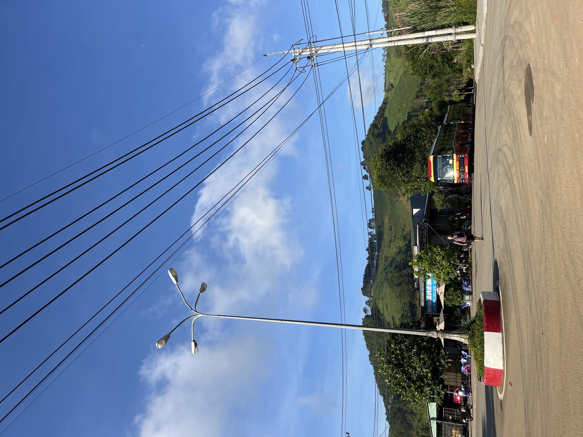 The image depicts a street scene in Bo Y, Vietnam, characterized by a clear blue sky with scattered white clouds. Multiple power lines stretch across the upper portion of the frame, supported by a tall lamppost with four lights in the left-center and a concrete utility pole with insulators on the right. In the foreground, a paved road forms a circular area with a traffic island in the center, which is decorated with a red and white border and filled with green foliage and yellow flowers.
A red and yellow bus is parked on the right side of the road. To the left of the bus, a building with outdoor seating, umbrellas, and chairs suggests a commercial establishment, possibly a restaurant or cafe. A person is walking away from the camera towards the right, carrying a dark backpack. A motorcycle is parked near the building on the left.
The background features a lush green hillside with terraced tea plantations, dotted with trees. The overall impression is a rural or semi-rural setting with infrastructure for transportation and commerce, set against a scenic natural landscape.