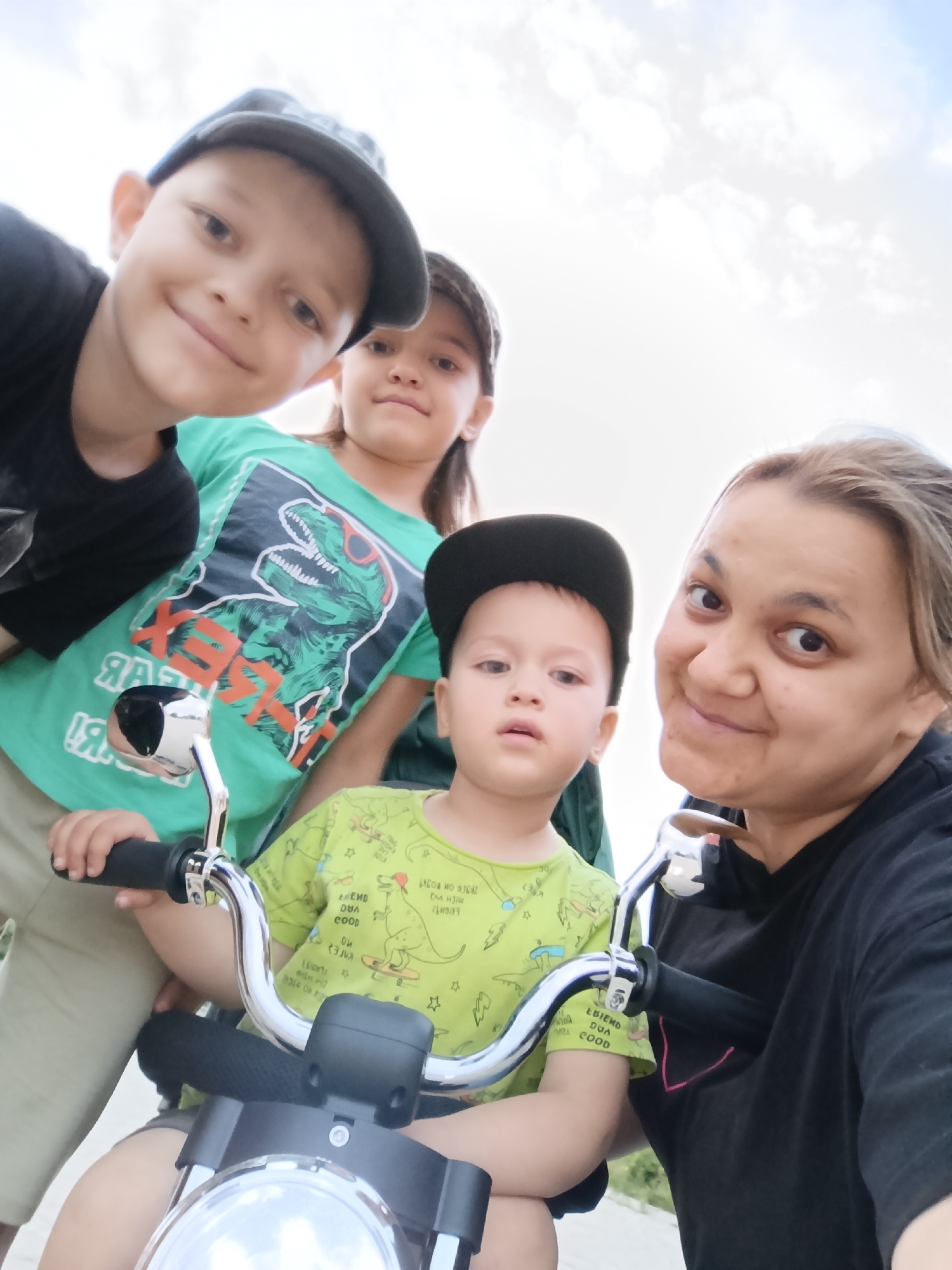 This is a selfie-style photo featuring four people, two adults and two children, with a bright, overcast sky in the background. The subjects appear to be outdoors, possibly in a park or recreational area, given the presence of a child's toy motorcycle in the foreground.
On the right side of the frame is an adult woman with light brown hair, smiling warmly at the camera. To her left and slightly behind her is a young boy wearing a light green t-shirt with a dinosaur print and a black baseball cap. He is positioned on the toy motorcycle, his hands gripping the handlebars. To the left of the boy and behind him is a young girl wearing a green t-shirt with a large dinosaur graphic on it. She is also looking towards the camera. In the front left, a boy wearing a dark t-shirt and a grey baseball cap is also visible, his face turned towards the camera and smiling.
The toy motorcycle has chrome handlebars and a headlight. Visible text on the children's t-shirts appears to be in a reversed, mirrored font, possibly a stylistic choice or due to the way the t-shirts are printed. The weather seems to be bright but cloudy, suggesting it could be daytime, possibly late morning or afternoon. The overall mood of the photo is cheerful and familial, with the individuals appearing to be enjoying an outing together. The presence of the toy motorcycle implies a playful or recreational activity.
