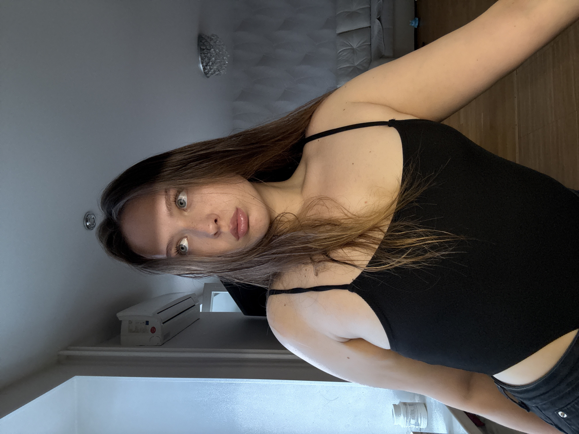 A young woman with long brown hair and blue eyes is captured in a candid shot, looking upwards and to her right with parted lips. She is wearing a black tank top and dark jeans. The background suggests an indoor setting, possibly a living room, with a modern chandelier visible on the ceiling and a sofa in the distance. A white air conditioning unit is mounted on the wall, and a window with vertical blinds is present on the left side of the frame, casting blue light onto the scene. This image was taken in Chaikovsky, Russia.