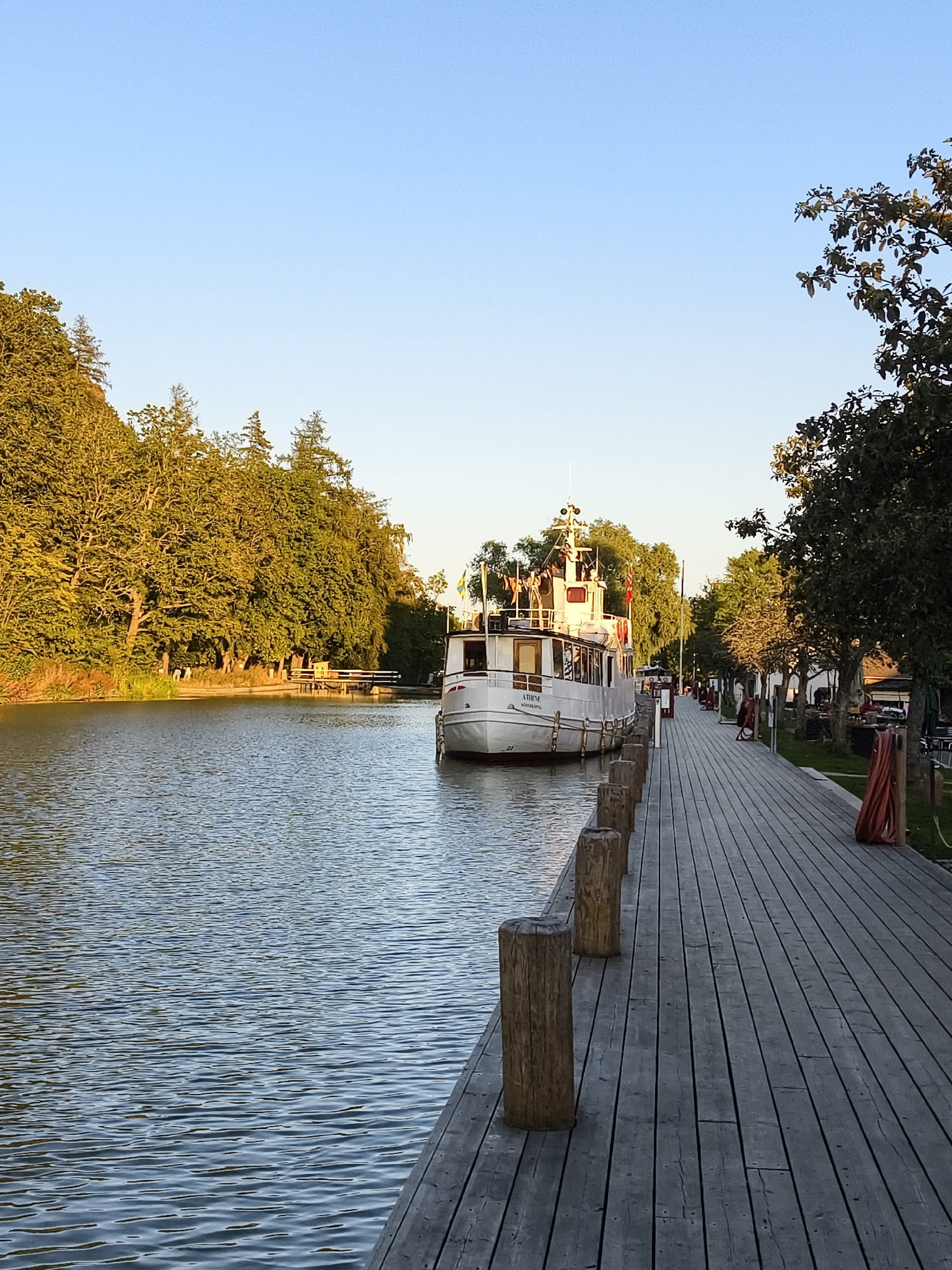 The image depicts a canal scene in Söderköping, Sweden. A white passenger boat named "ATHENE" is moored alongside a wooden boardwalk. The boardwalk runs along the right side of the canal, with several bollards placed at intervals. Trees line the left bank of the canal, with lush green foliage under a clear blue sky. The water in the canal is calm with gentle ripples. Several people are visible on the boardwalk, some walking and others standing. Buildings and more trees are present in the background on the right side of the canal. The overall impression is a peaceful waterside setting, likely during daylight hours given the clear sky and illumination.